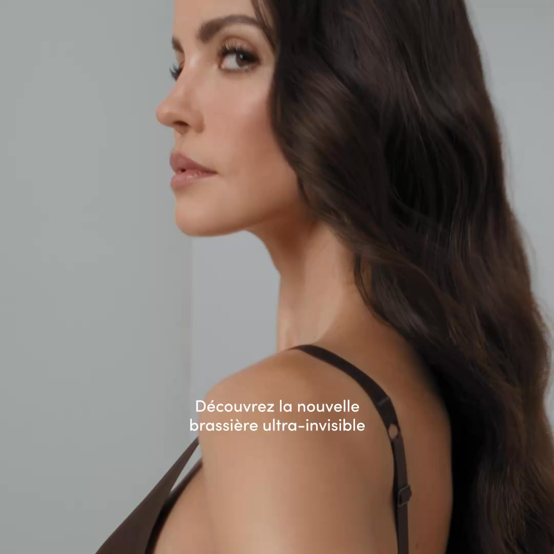 A close-up, profile shot of a woman with dark, wavy hair looking over her shoulder. She is wearing a dark-colored bra. White text is overlaid on the right side of the image, stating "Découvrez la nouvelle brassière Aria ultra-invisible. Sans armatures, sans coutures et incroyablement douce. Elle épouse naturellement votre silhouette pour un confort absolu." The overall aesthetic is clean and sophisticated, focusing on the product's features and the model's appearance.