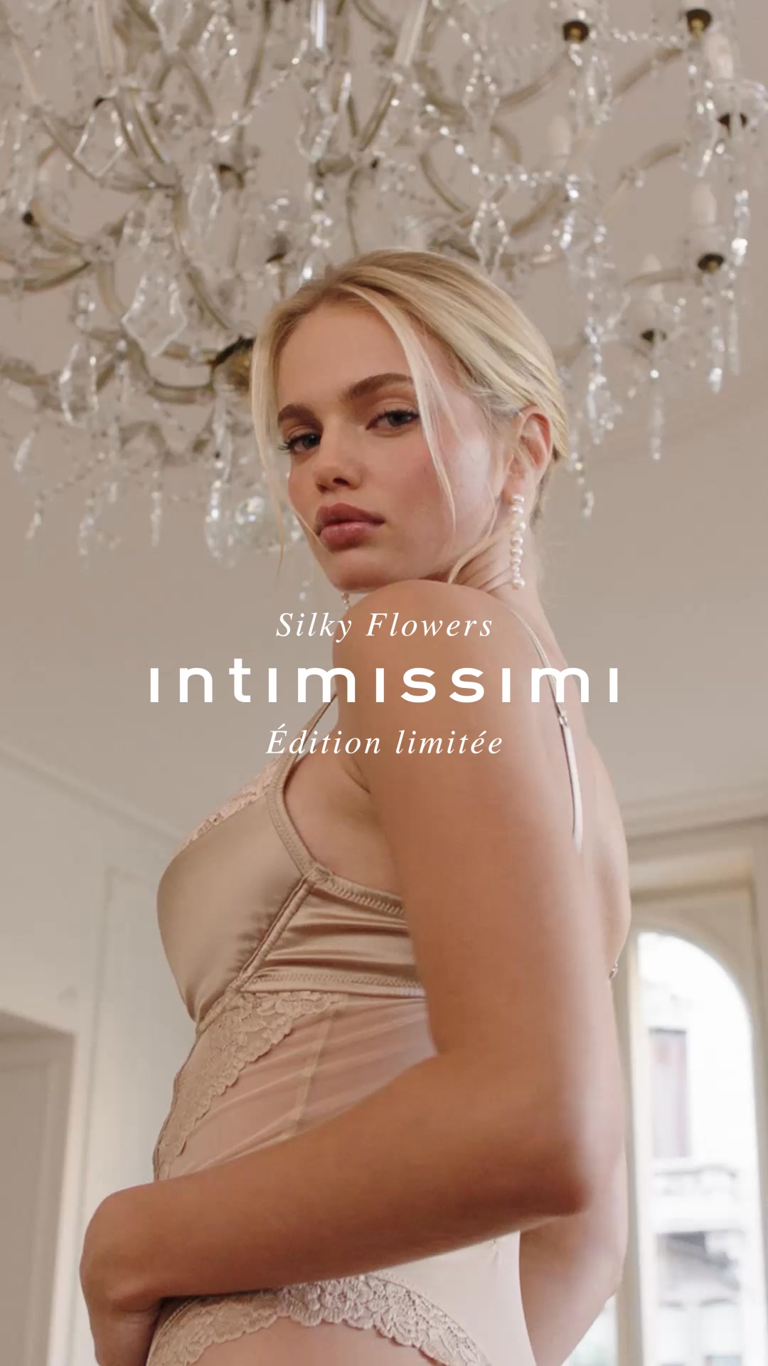 A blonde model with fair skin and blue eyes is shown from the chest up, looking over her shoulder towards the camera. She is wearing a champagne-colored satin slip with delicate lace trim. Above her, a crystal chandelier is visible. Text overlays on the image read "Silky Flowers" and "Intimissimi" in a large, elegant font, followed by "Édition limitée" in a smaller font below.