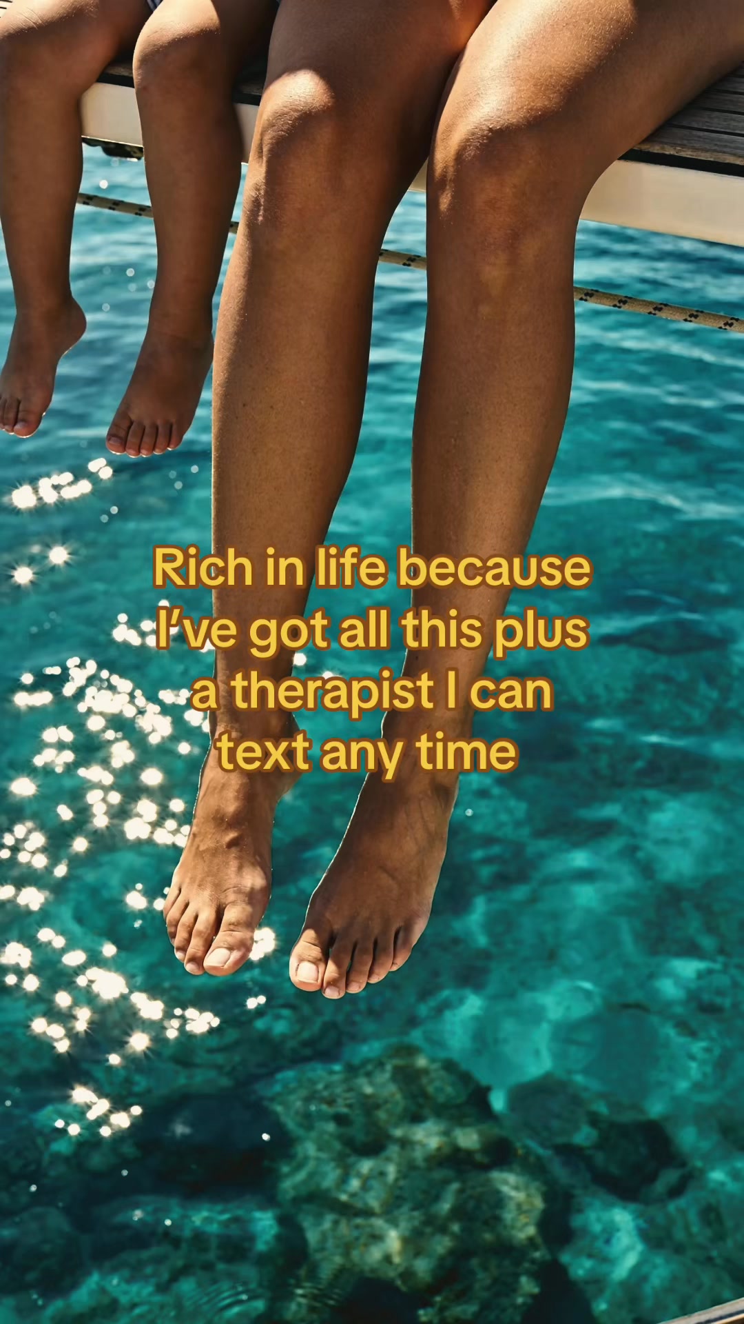 The image shows the lower legs and feet of two people sitting on the edge of a boat, with their feet dangling over the bright blue, sun-dappled water. Yellow text is overlaid on the image, reading: "Rich in life because I've got all this plus a therapist I can text any time." The creative focuses on the feeling of contentment and the accessibility of mental health support.