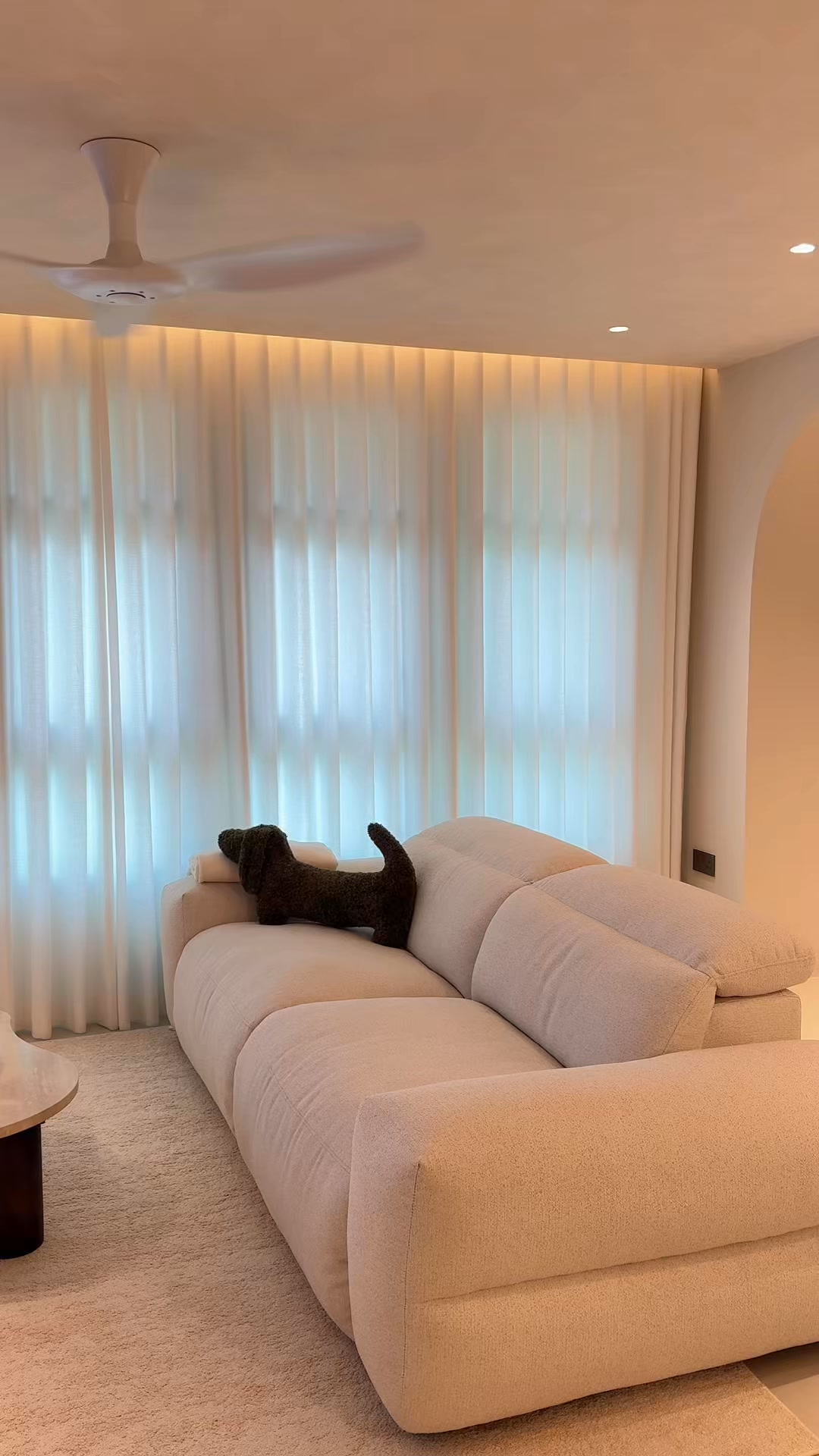 A plush, cream-colored sectional sofa is shown in a well-lit, modern living room. A dark green, dachshund-shaped pillow rests on the sofa. The sofa is positioned in front of sheer, floor-to-ceiling curtains that diffuse natural light. The overall aesthetic is minimalist and comfortable, highlighting the sofa as a central piece of furniture for relaxation and everyday living.