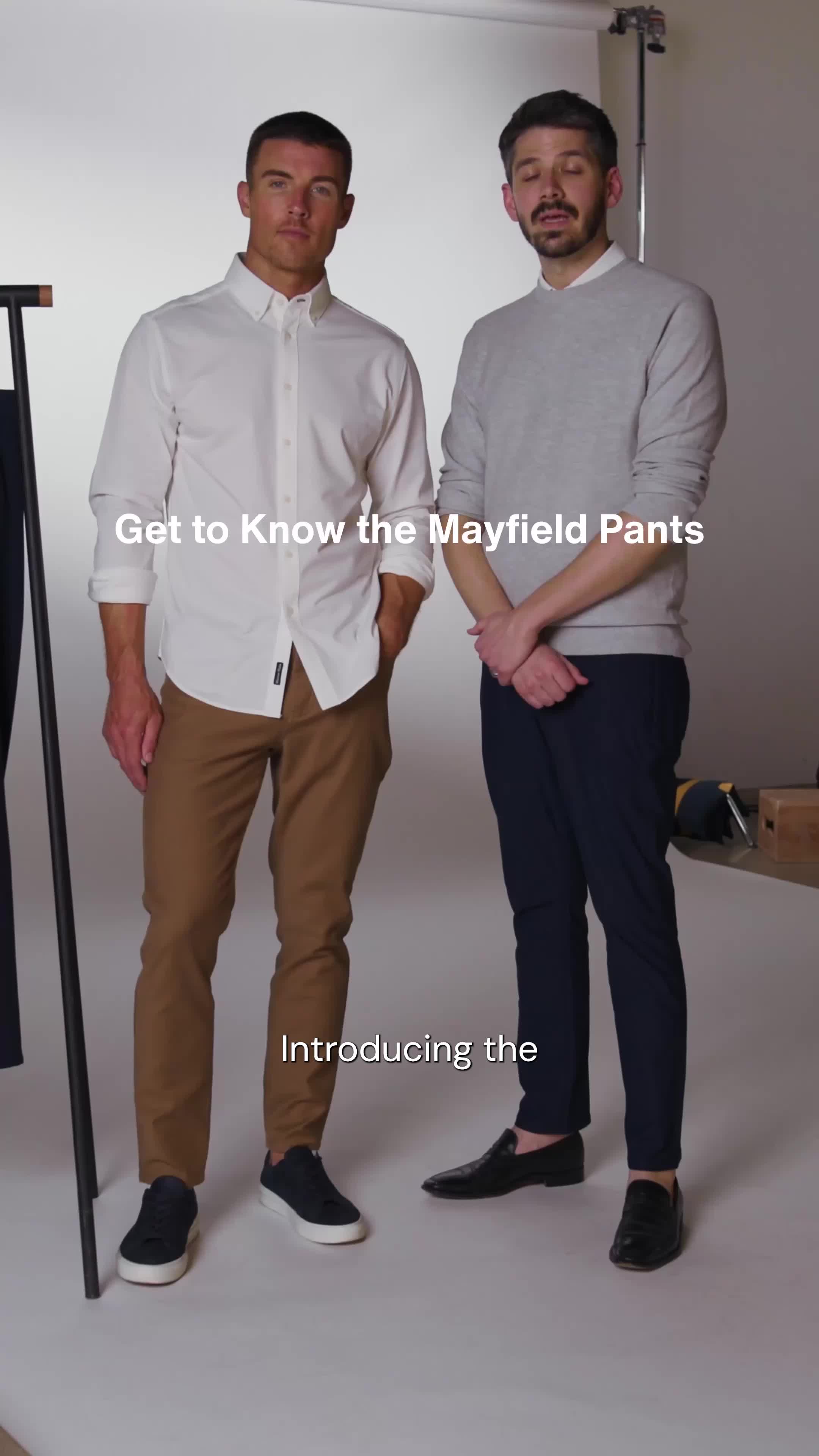 Two men are standing side-by-side in a studio setting. The man on the left wears a white button-down shirt and brown pants, while the man on the right wears a grey sweater and navy pants. White text overlaid on the image reads "Get to Know the Mayfield Pants" and "Introducing the". The overall aesthetic is clean and professional, highlighting the apparel.