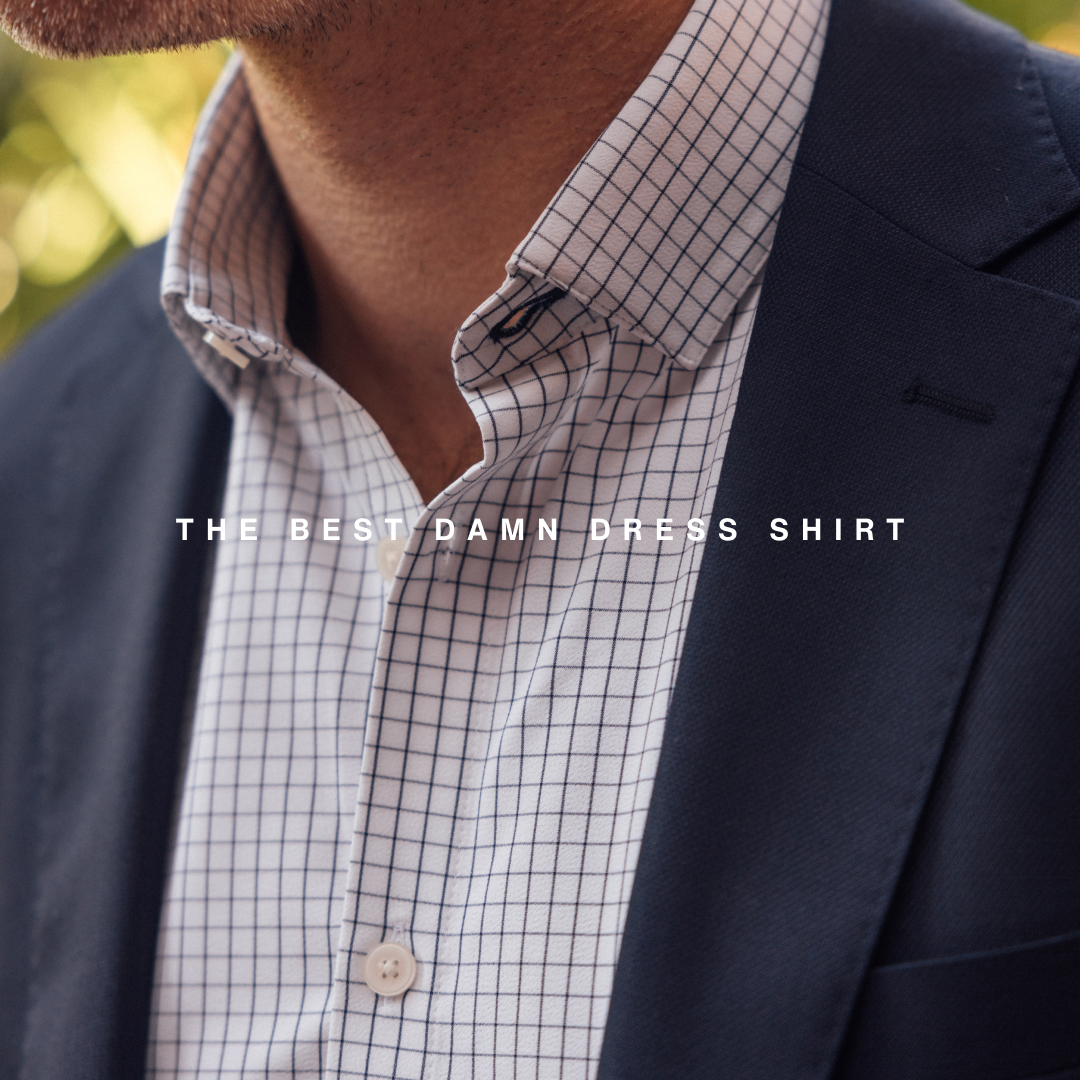 A close-up shot focuses on the chest area of a man wearing a navy blue suit jacket over a white dress shirt with a subtle blue checkered pattern. The shirt collar is visible, along with a button. White text is overlaid on the image, centered horizontally, stating "THE BEST DAMN DRESS SHIRT". The background is softly blurred, suggesting an outdoor setting with greenery.