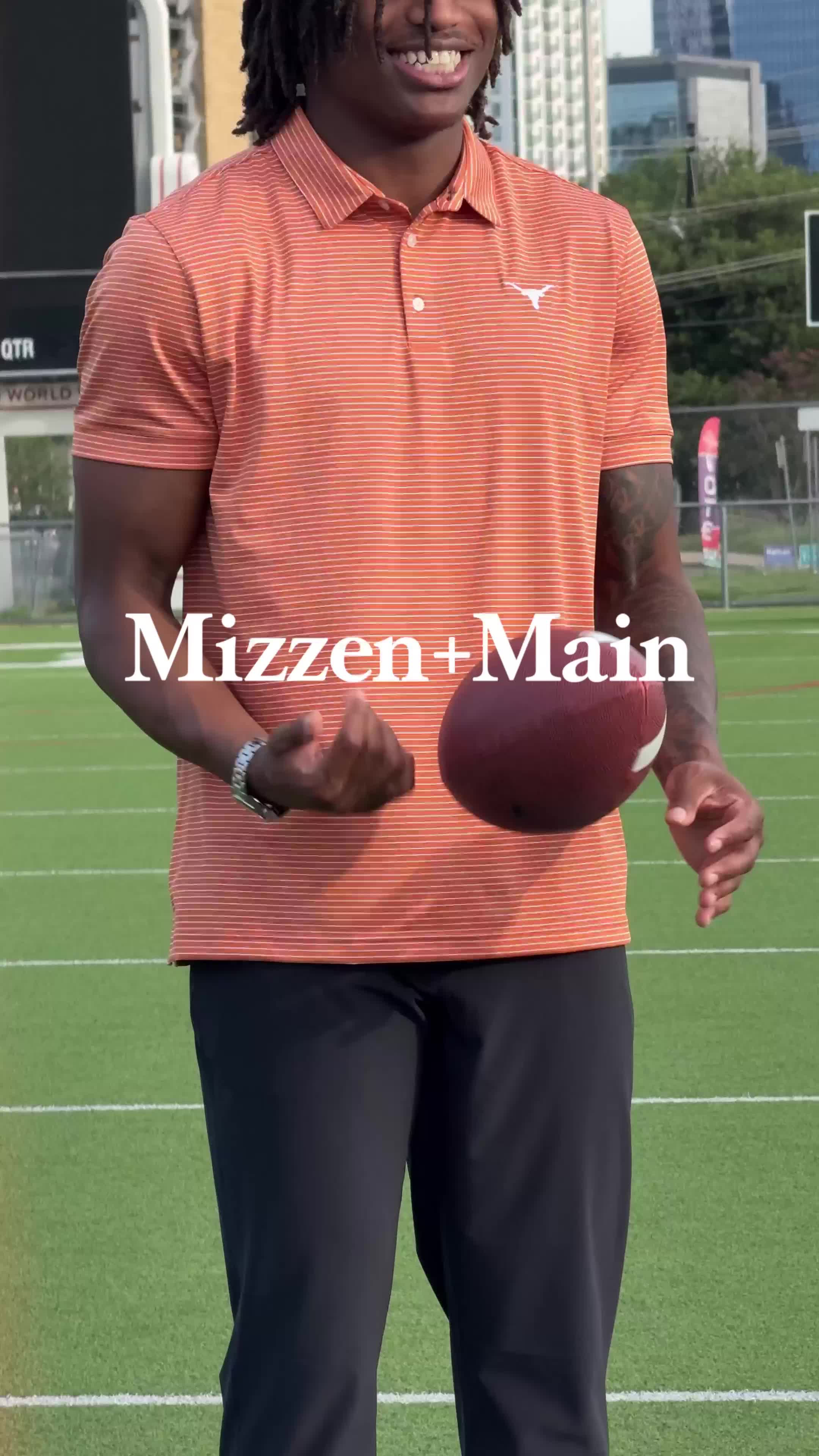 A man wearing an orange and white striped polo shirt stands on a green field, holding a football. Text overlay reads "Mizzen+Main". The man is smiling and looking towards the camera. The background shows a blurred cityscape and stadium elements. The copy invites the viewer to see what a UT Wide Receiver wears off the field and purchase similar styles.