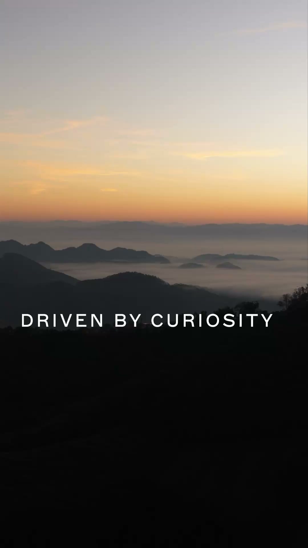 The image shows a serene mountain landscape at sunrise, with layers of mist settling in the valleys between the peaks. The sky transitions from a soft orange glow near the horizon to a pale blue overhead. White text overlaid on the lower half of the image reads "DRIVEN BY CURIOSITY". The overall mood is peaceful and contemplative, evoking a sense of exploration and wonder.