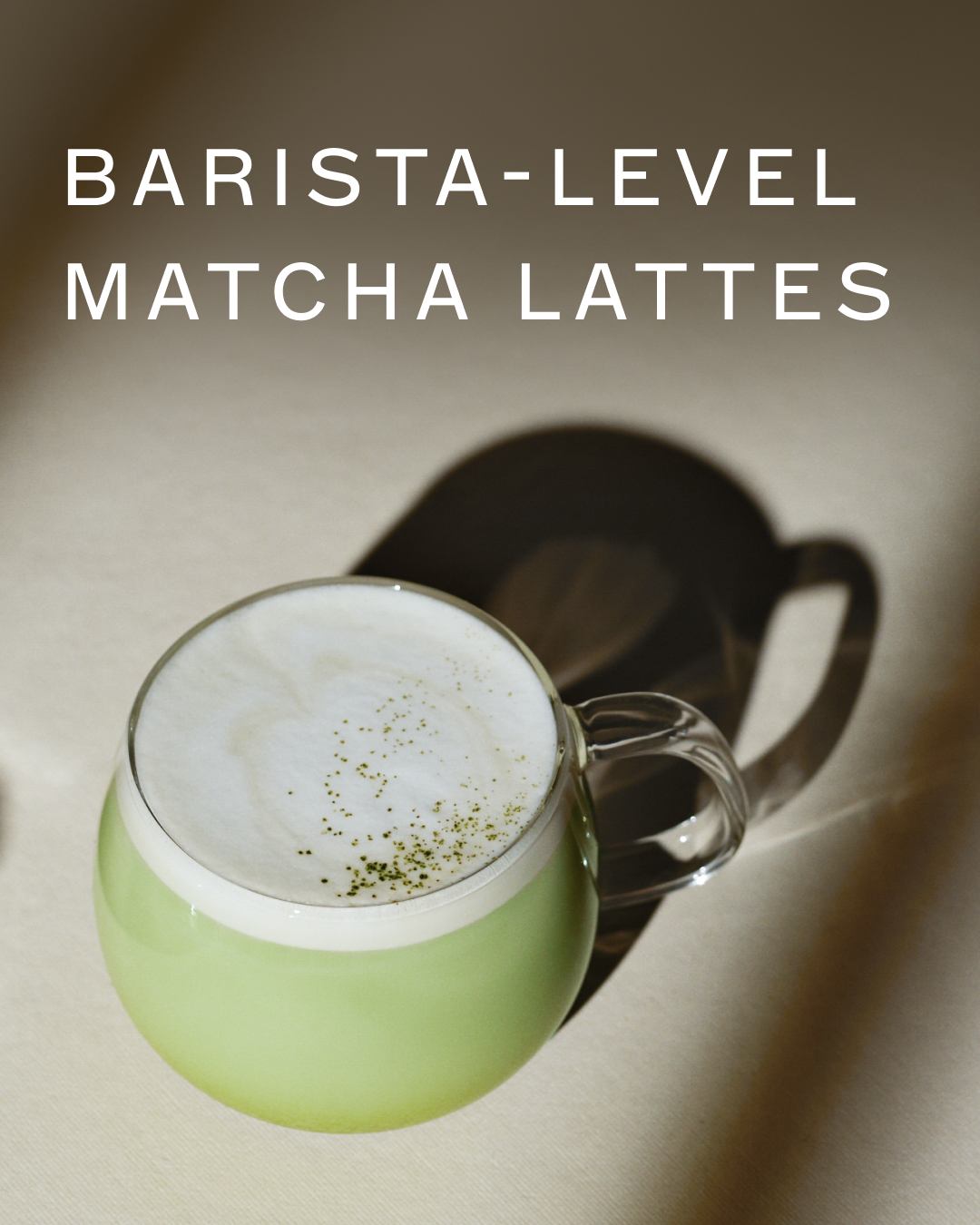 A close-up shot of a light green matcha latte in a clear glass mug, topped with white foam and a sprinkle of matcha powder. White text overlayed on the top of the image reads "BARISTA-LEVEL MATCHA LATTES". The lighting creates a soft shadow on the right side of the mug.