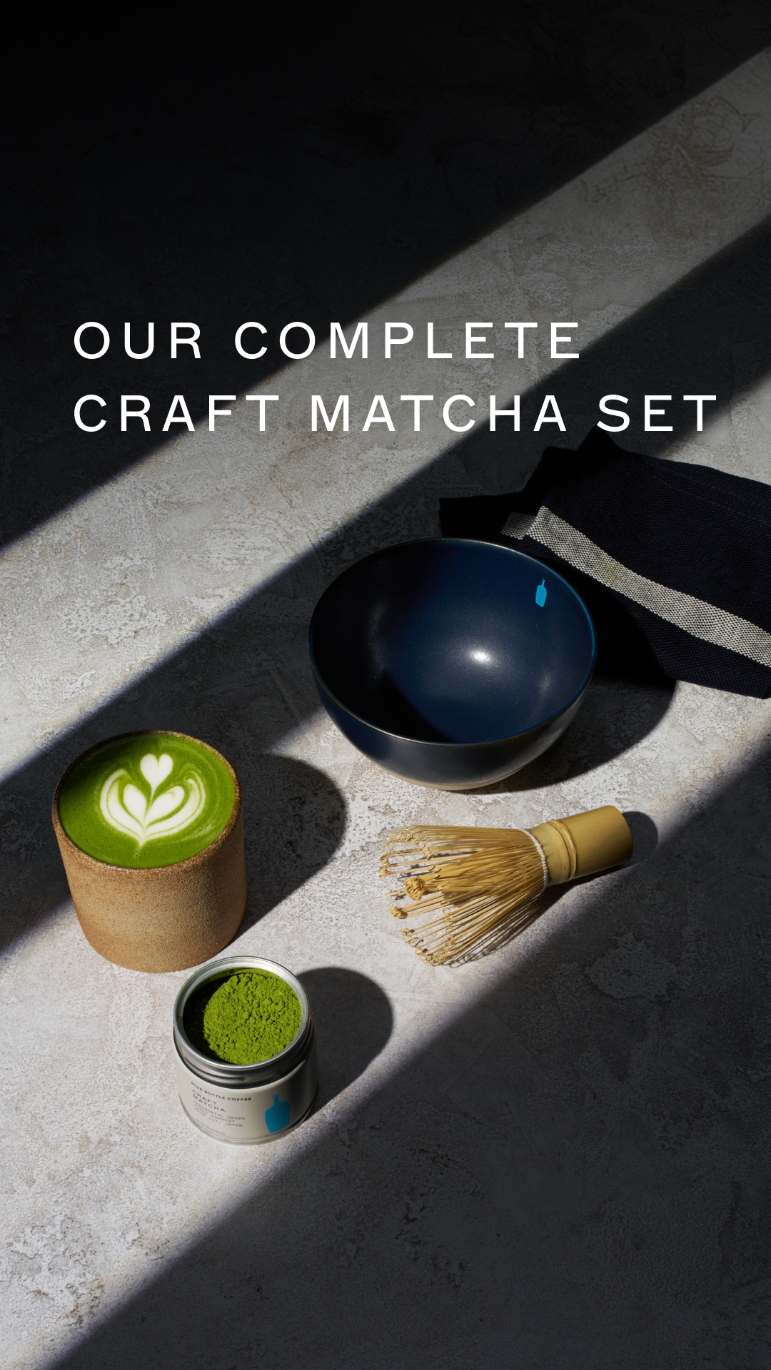 A flat lay image shows a matcha set on a textured surface, bathed in dramatic light and shadow. The set includes a ceramic cup with latte art, a tin of matcha powder, a dark blue bowl, and a bamboo whisk. White text overlaid on the image reads "OUR COMPLETE CRAFT MATCHA SET", directly addressing the viewer and highlighting the product.