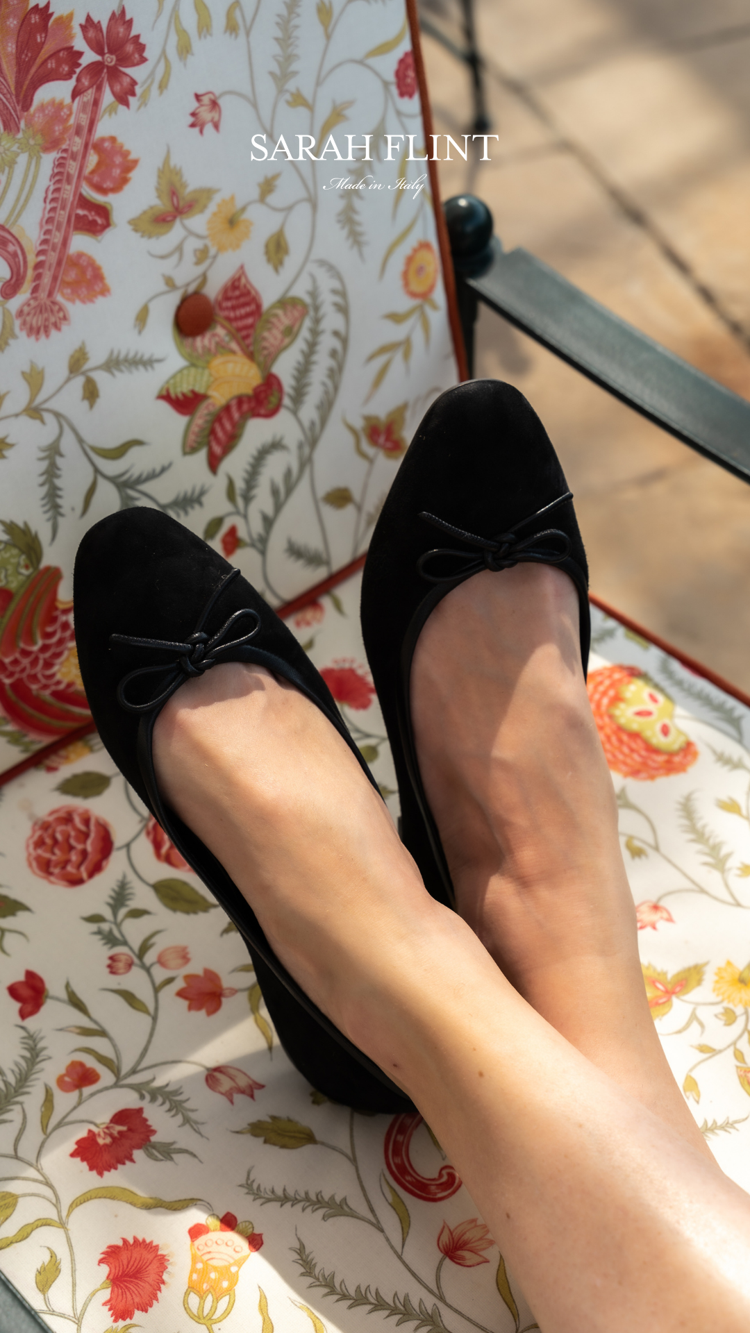 A close-up shot shows a person's feet wearing black suede ballet flats with bows. The feet are crossed and resting on a floral-patterned chair. The brand name "SARAH FLINT" and "Made in Italy" are displayed in white text at the top of the image. The text overlay is a five-star review praising the comfort of the shoes.
