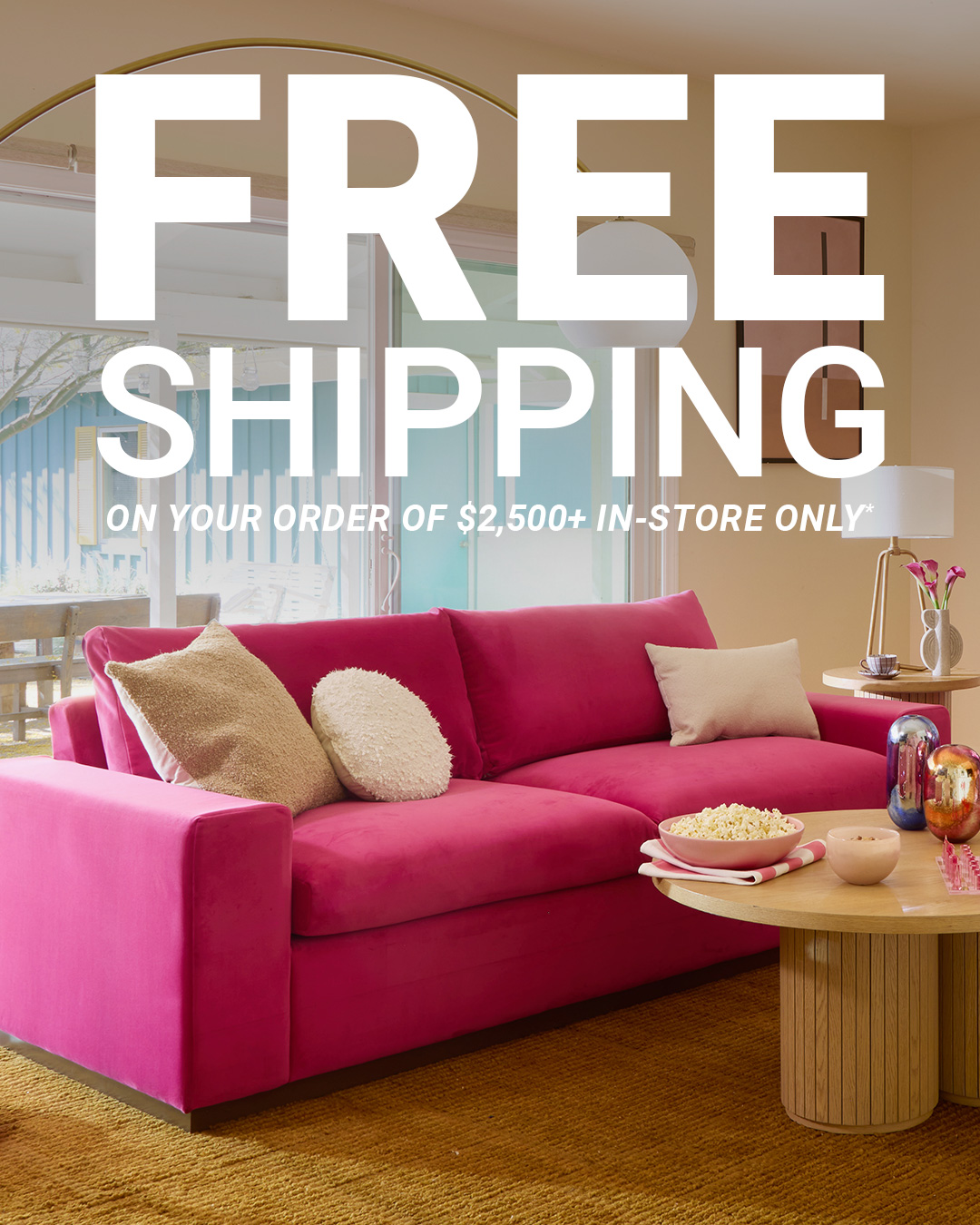 A bright, modern living room features a plush, fuchsia sectional sofa. Large white text overlays the image, proclaiming "FREE SHIPPING" across the top two-thirds of the frame. Below this, smaller text reads "ON YOUR ORDER OF $2,500+ IN-STORE ONLY*." The scene is set with decorative pillows on the sofa and a coffee table with a bowl of popcorn and decorative vases.