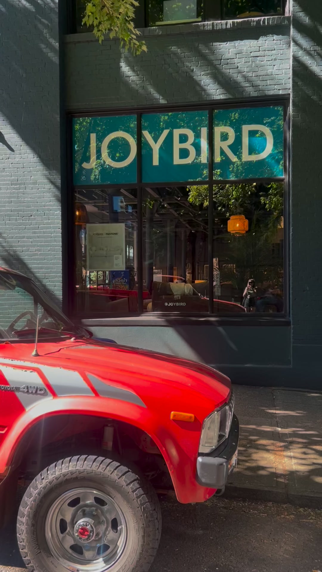 A bright red vintage Toyota 4WD truck is parked on a sunny street in front of a building with large windows. The window displays the word "JOYBIRD" in large, white, sans-serif letters against a teal background. Inside the store, furniture and decor are visible. The text overlay on the image reads: "Take pieces for a sit-test before you buy at one of our nationwide showrooms and save up to 40% off during Joybird's Labor Day Preview Sale."