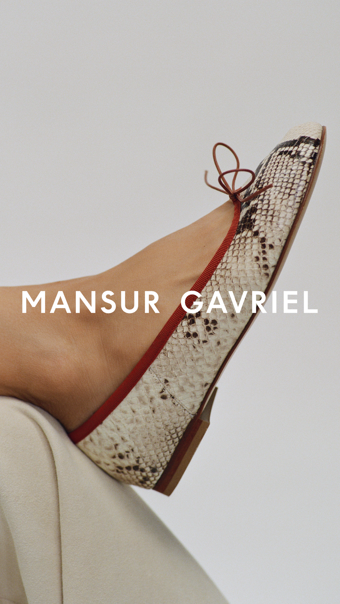 A close-up shot focuses on the side of a woman's foot wearing a beige, square-toed ballet flat with a snakeskin pattern. The shoe has a thin red trim and a small brown bow at the toe. The brand name "MANSUR GAVRIEL" is displayed in large white sans-serif font across the image, overlaying the foot and shoe. The background is a plain, light grey, and the lower left corner shows a hint of beige fabric, likely clothing.