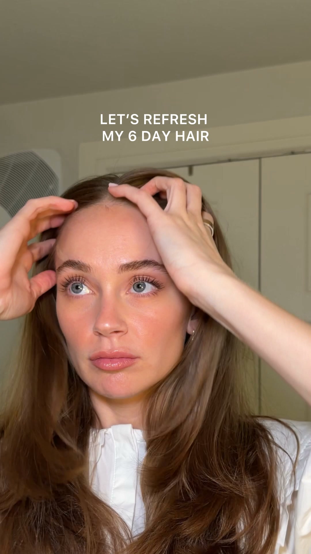 A close-up shot of a woman with light brown hair, looking directly at the camera. She is touching her hair and forehead with both hands. White text overlaid on the top of the image reads "LET'S REFRESH MY 6 DAY HAIR". The woman is wearing a white collared shirt. The background appears to be a room with a vent on the left and a closet door on the right.