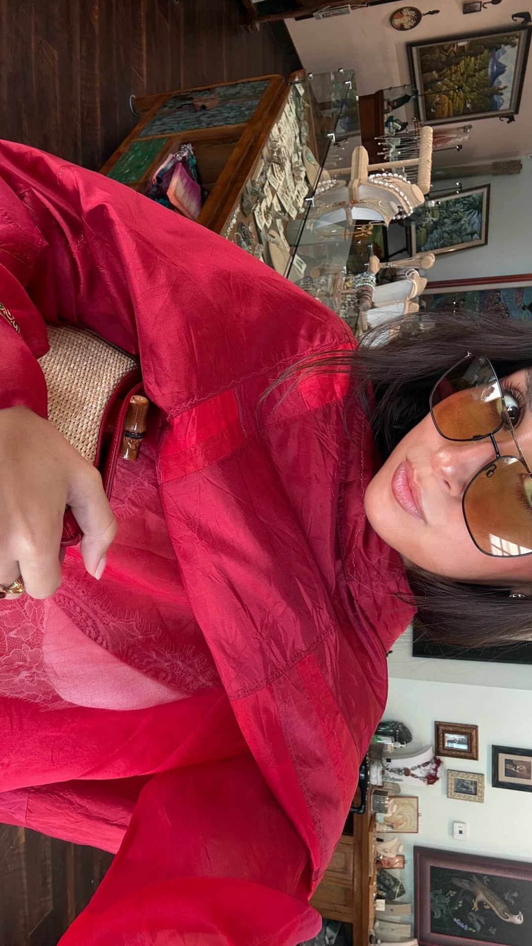 A woman with dark hair and large sunglasses is wearing a bright red jacket and holding a straw bag. She is looking towards the camera with a slight smile. The background appears to be an antique shop with display cases filled with jewelry and artwork on the walls. Text overlays the image, stating 'Innovation meets dry shampoo skeptics for an unexpected match made in voluminous bliss @livingproofinc #LivingProofPartner available now @sephora !'