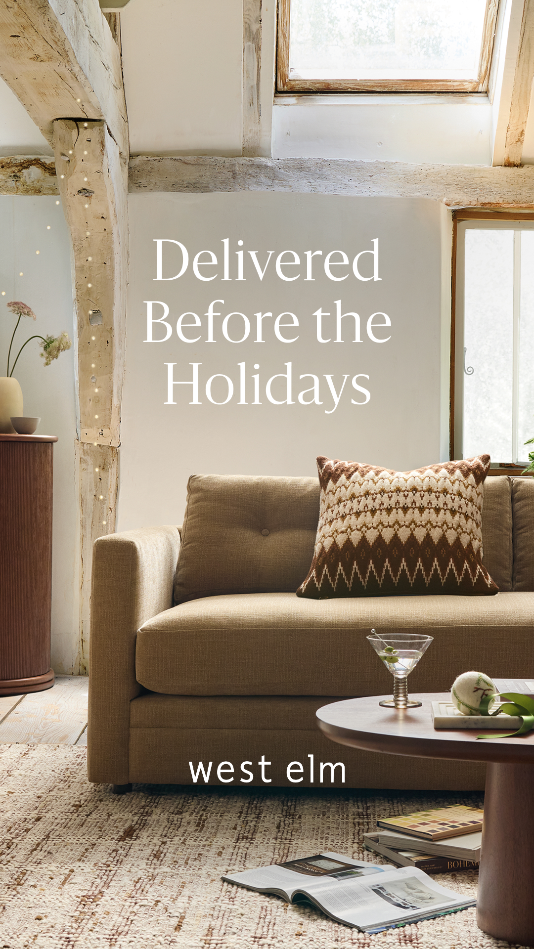 A cozy living room scene is depicted with a sofa, a coffee table, and a rug. The sofa has a decorative pillow with a geometric pattern. On the coffee table, there is a martini glass with olives and a small decorative item. The text "Delivered Before the Holidays" is overlaid in large, elegant font at the top of the image. Below the sofa, the West Elm logo is displayed. The overall aesthetic suggests a comfortable and festive holiday setting, emphasizing timely delivery for seasonal preparations.