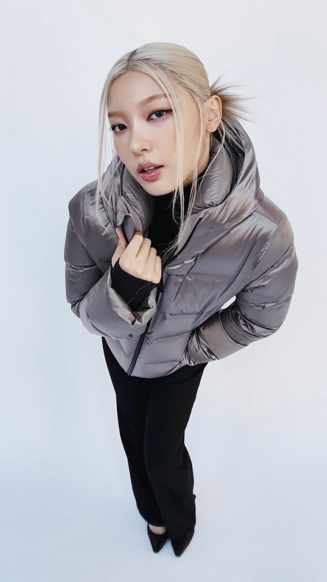 A young woman with blonde hair tied back in a ponytail, wearing a grey puffer jacket over a black turtleneck and black pants, is shown from a high angle against a white background. She is looking towards the camera with her hands near her neck. The text '@yoonchae likes her AlpenGlaze™ options.' is overlaid on the image.