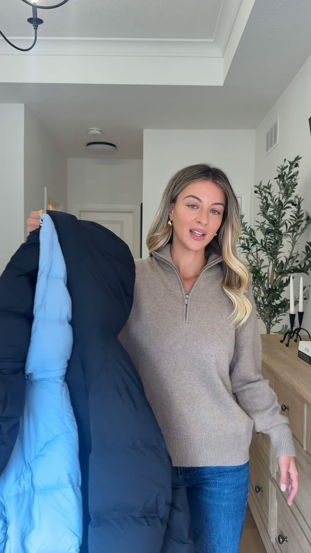 A woman stands in a hallway, holding up a dark blue puffer jacket with a light blue lining. She is wearing a beige quarter-zip sweater and blue jeans, and has blonde wavy hair and gold hoop earrings. Text overlaying the image reads: "For superior warmth, wear The Super Puff™ like @melvalentini." The background shows a dresser with decorative items and a plant.