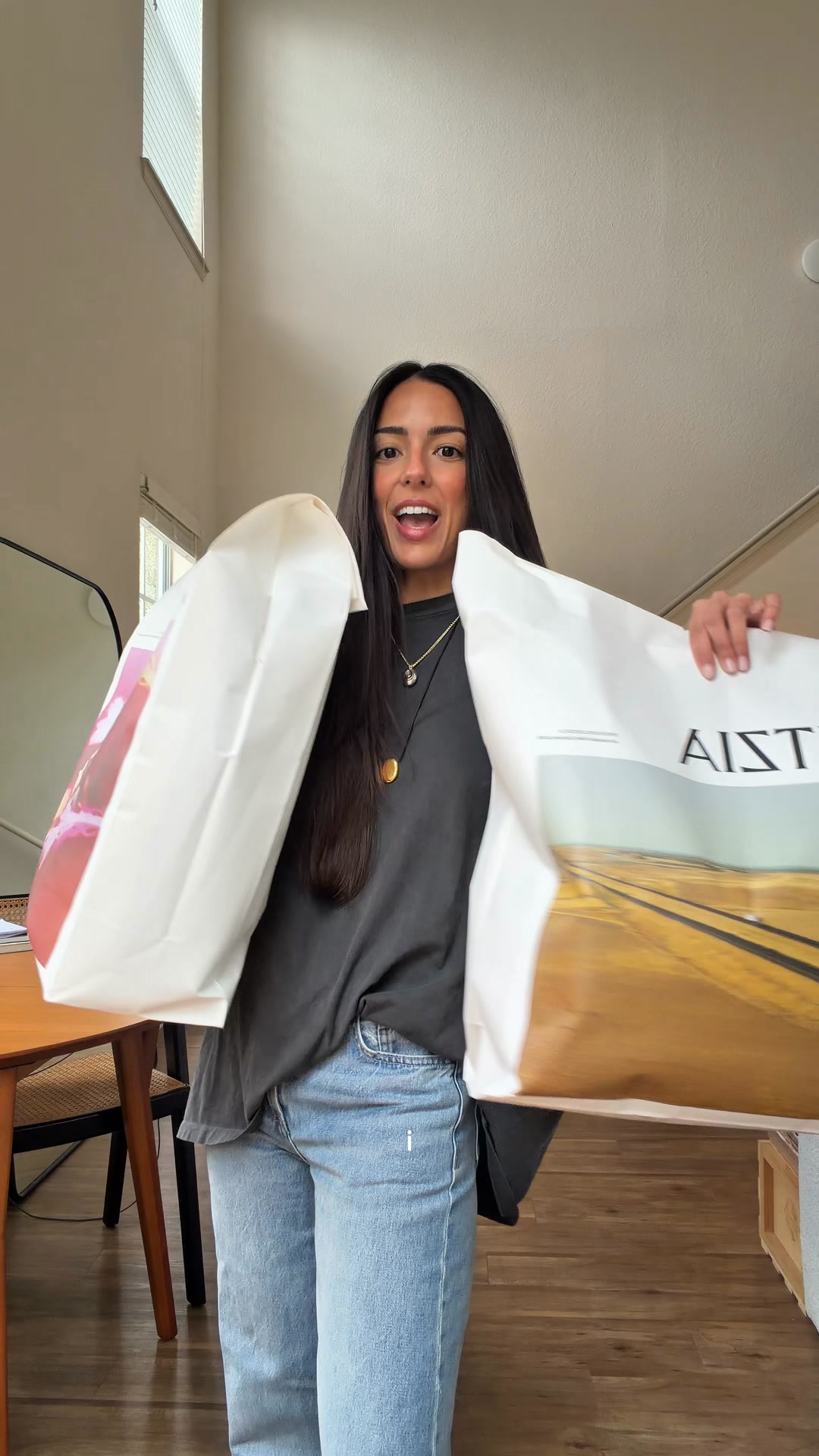 A woman with long dark hair and a wide smile holds up two large shopping bags, one in each hand. She is wearing a dark grey t-shirt and light blue jeans. The bags are white and appear to contain items from a store. The background is a bright, airy room with wooden floors and a tall wall. Text overlaying the image reads "Aritzia must-haves. As seen on @taynnabrito".