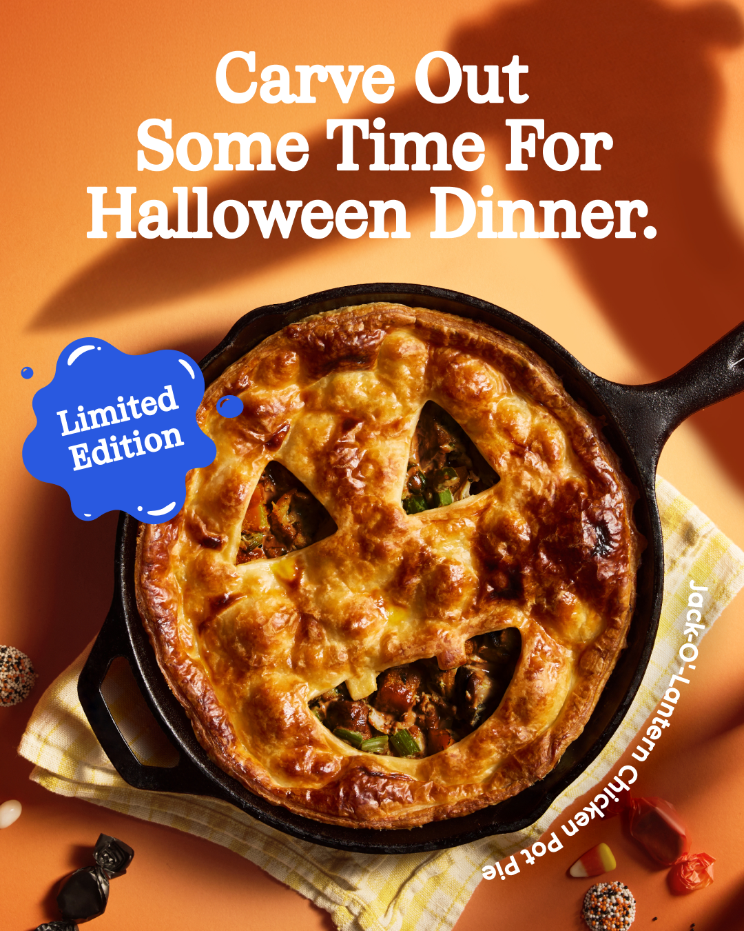 A Halloween-themed chicken pot pie, shaped like a jack-o'-lantern, is presented in a cast-iron skillet on a yellow checkered cloth. The pie has a golden-brown crust with a carved jack-o'-lantern face. Text overlays read "Carve Out Some Time For Halloween Dinner." and "Limited Edition" in a blue speech bubble. The bottom right corner has text that reads "Jack-O'-Lantern Chicken Pot Pie" and some Halloween candies are scattered around the pie.