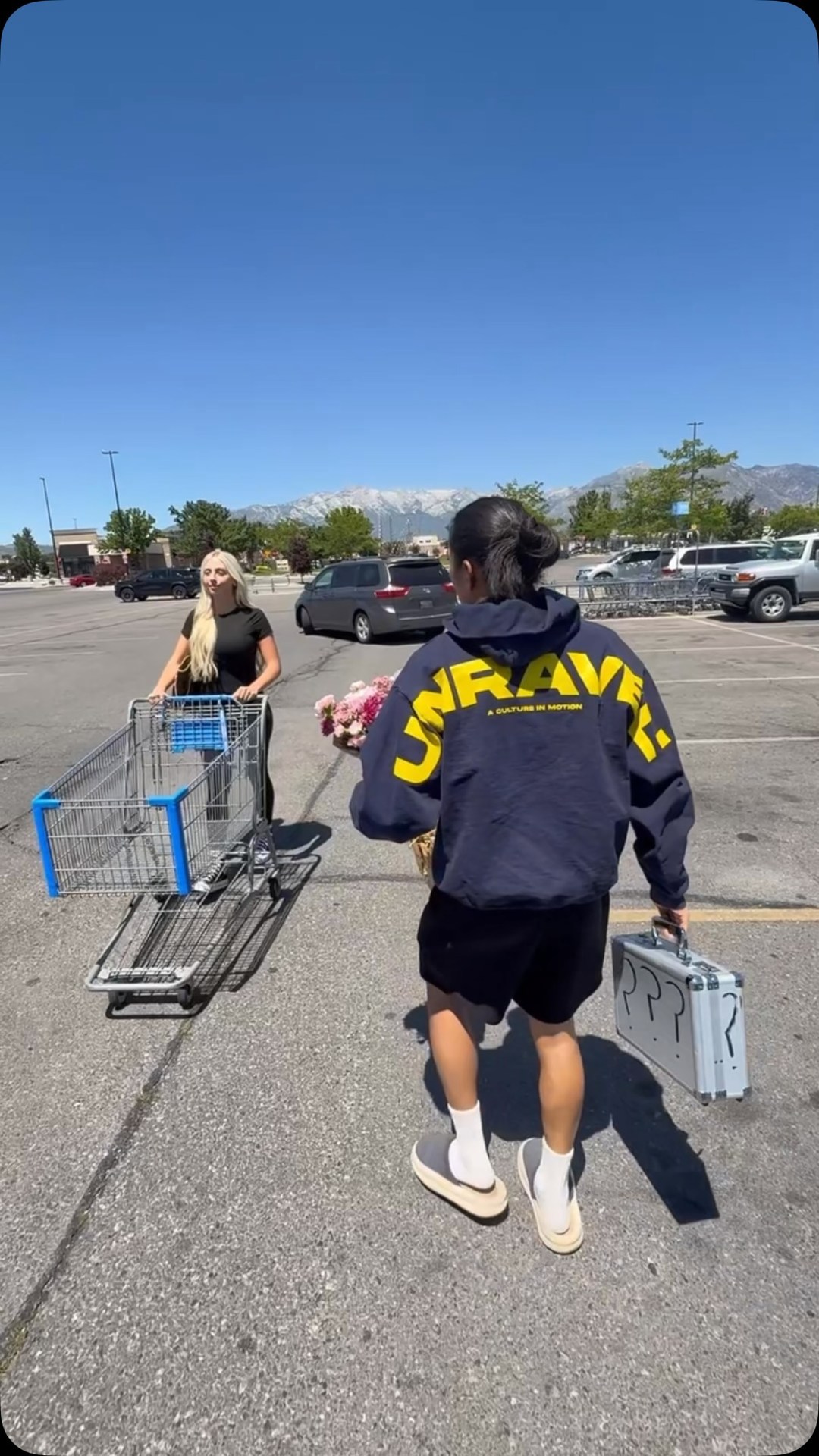 A blonde woman with a shopping cart and a person with dark hair wearing a navy hoodie with yellow lettering that says "UNRAVEL" stand in a parking lot. The person in the hoodie is holding a bouquet of flowers and a silver briefcase with question marks on it. The blonde woman is looking at the person in the hoodie. The sky is clear and blue with mountains in the background. The text on the creative says "I handcuffed myself to this girl for 24 hours for the ultimate financial mystery date with @acorns".
