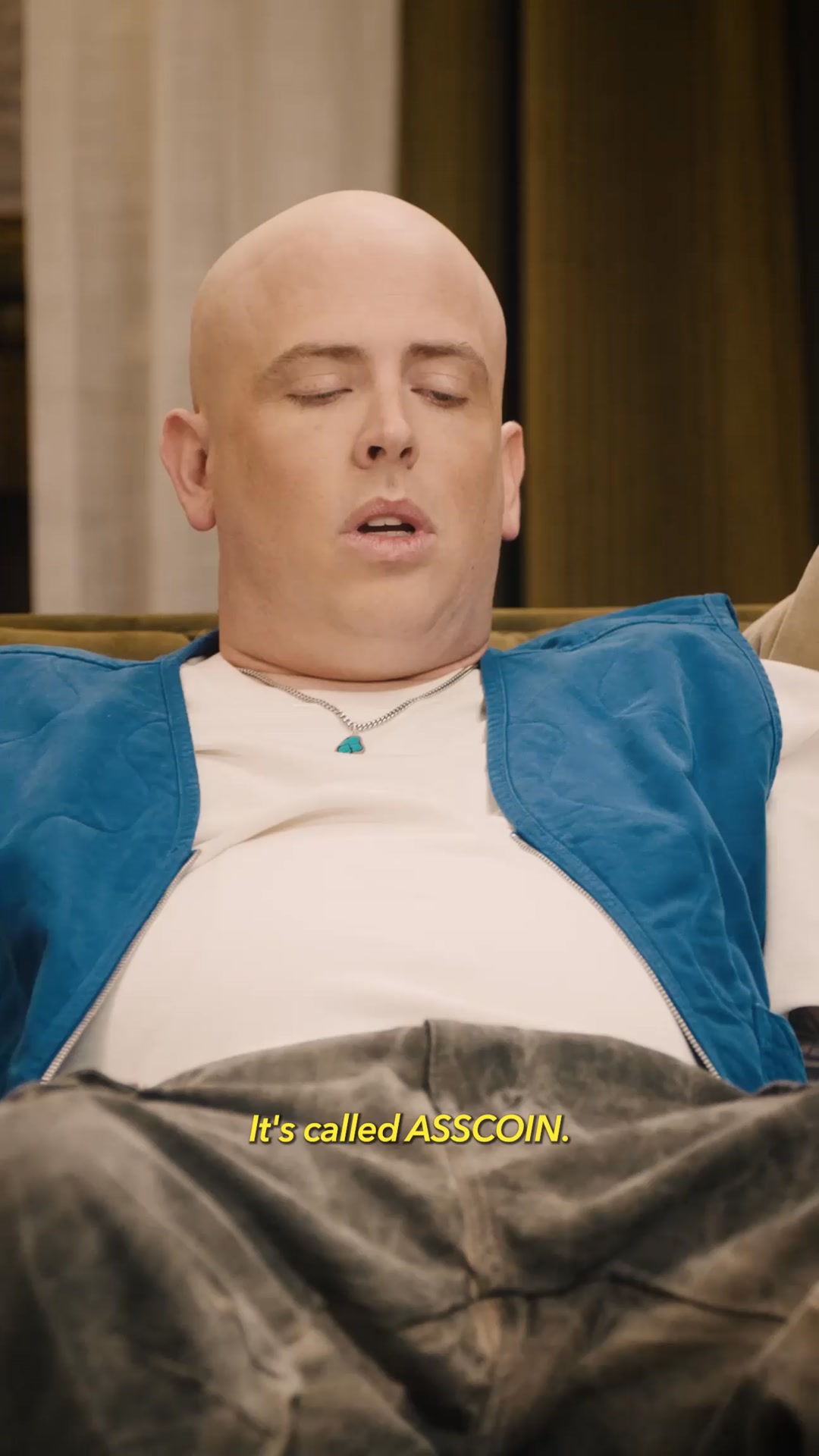 A close-up shot of a bald person with their eyes closed, wearing a blue vest over a white t-shirt and a silver necklace with a turquoise pendant. The person is sitting on a couch. Yellow text overlaid on the bottom of the image reads "It's called ASSCOIN."