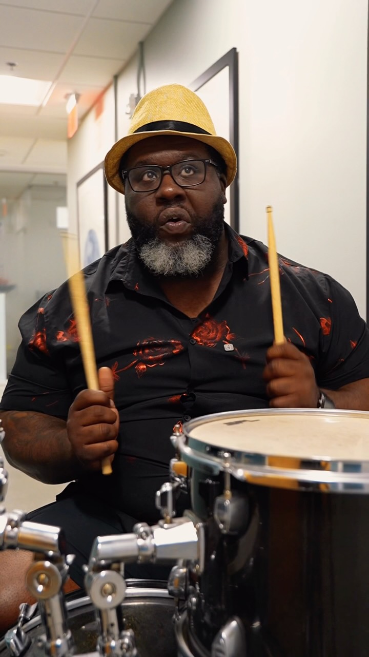A man wearing a yellow fedora and a black shirt with red floral patterns is playing the drums. He is looking up and to the left with an open mouth, holding drumsticks. The text "Nobody told Pops how easy investing is now, so I had to. Every swipe rounds up and goes into your portfolio #AcornsPartner #GrowYourOak" is overlaid on the image.