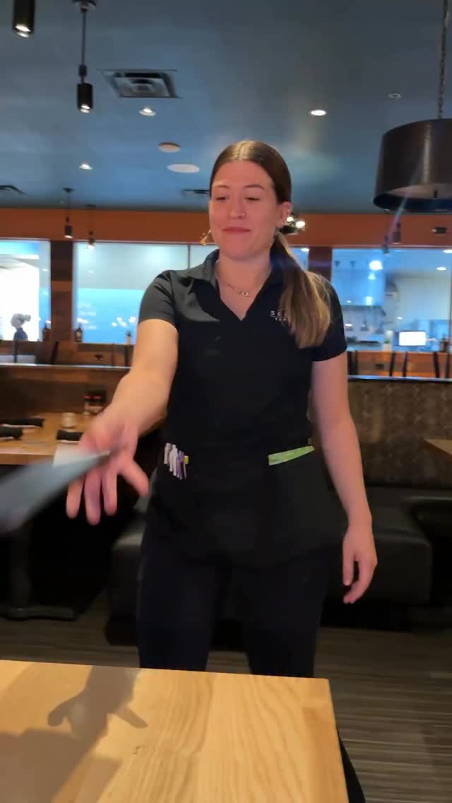 A woman in a black polo shirt and apron stands in a restaurant, holding a menu. She is smiling and looking towards the camera as she extends her arm, appearing to hand the menu to the viewer. The background shows tables and booths typical of a dining establishment. The text "Dinners on me!" is overlaid on the image.