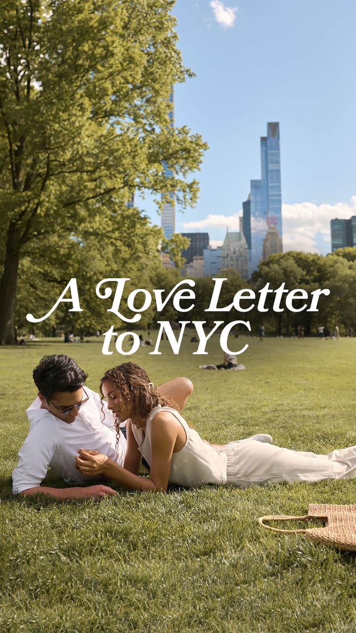 A couple is lying on the grass in a park with a city skyline in the background. The text "A Love Letter to NYC" is overlaid on the image in a script font. The scene is bright and sunny, with trees and other people in the park. The overall impression is one of romance and urban enjoyment.