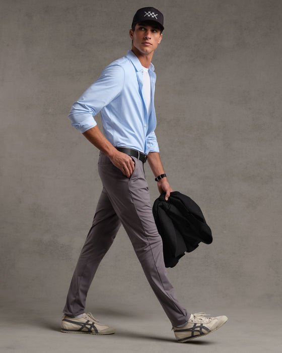 A male model walks forward, looking to his right, against a plain grey background. He wears a light blue button-down shirt, a white t-shirt underneath, grey pants, a black belt, and a black baseball cap. He holds a black jacket in his left hand. Text overlays the image: "Designed to move. Made with your comfort in mind. Take 15% off your first order and feel the Rhone difference."