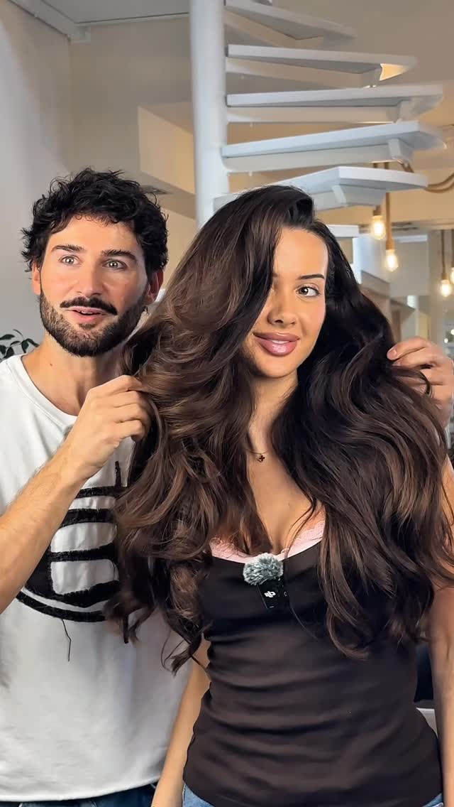 A man with dark curly hair and a beard, wearing a white t-shirt, is standing behind a woman with long, voluminous brown hair. He is gently touching her hair. The woman is looking towards the camera with a smile. Text overlays the image, highlighting a "Bombshell Look" created with a specific product and tagging the brand and influencer.