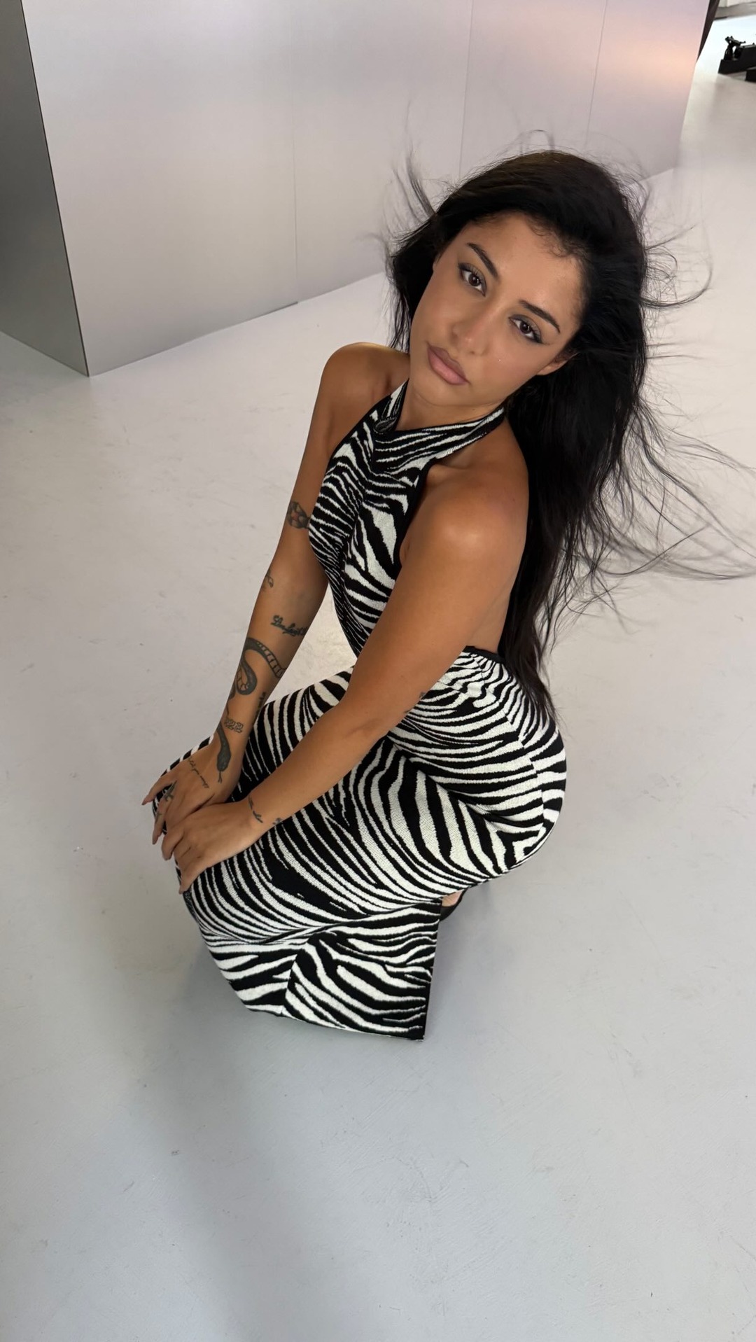 A woman with long, dark hair is kneeling on a white floor, looking towards the camera. She is wearing a black and white zebra-print halter dress. Her hair is blowing to the side, creating a dynamic effect. Text overlays the image, stating "#ADV @nssgclub @colorwowhair ho trovato il cheat code per i perfetti glass hair ✨ #lorealitaliatribe #dreamcoat #haircare".
