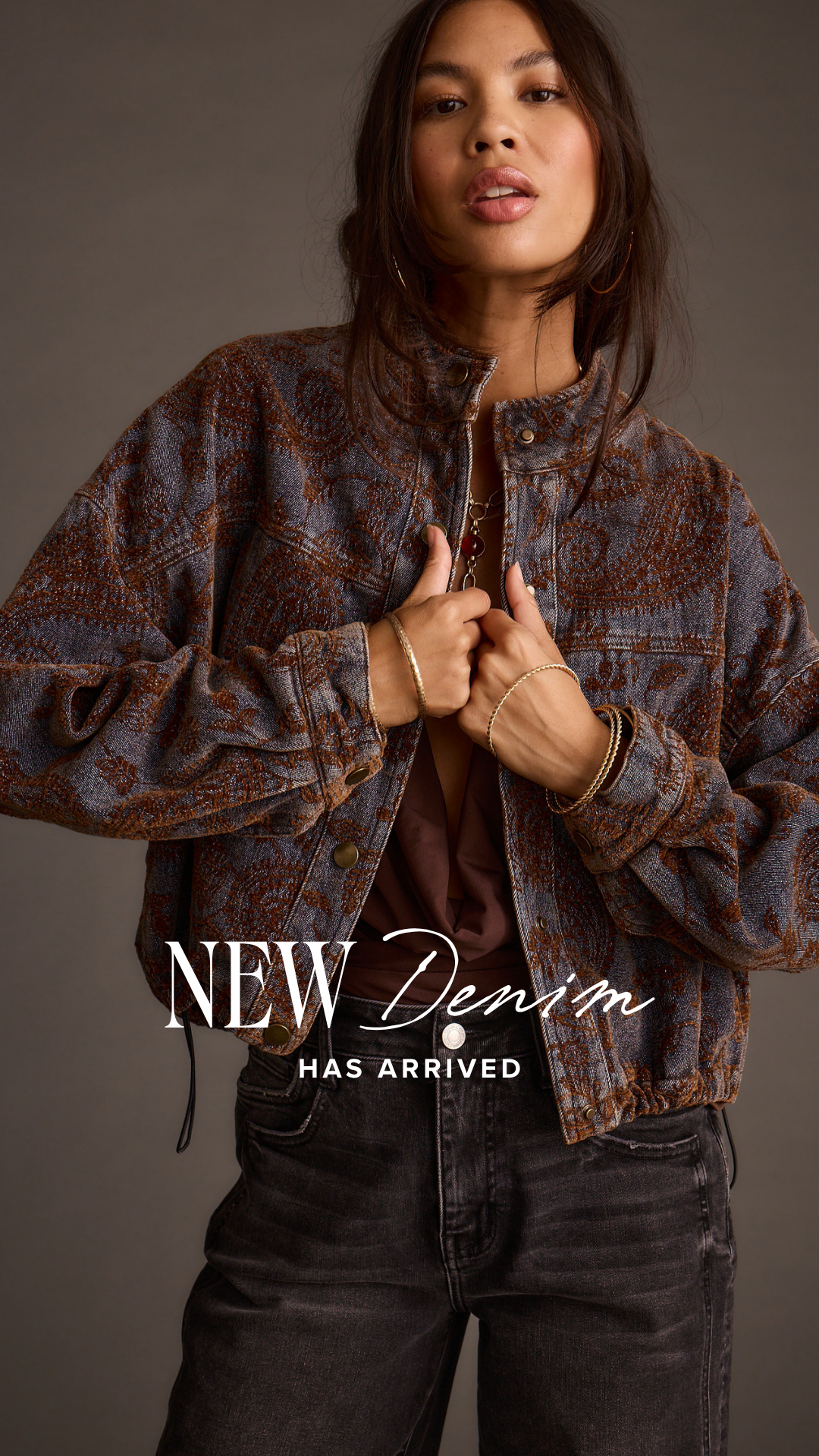 A woman is shown from the chest up, wearing a patterned denim jacket and dark wash jeans. She is holding the lapels of her jacket with both hands, looking towards the camera with her mouth slightly open. White text overlays the bottom half of the image, reading "NEW Denim" in a large, stylized font, with "HAS ARRIVED" in a smaller, simpler font below it. The background is a plain, dark grey.