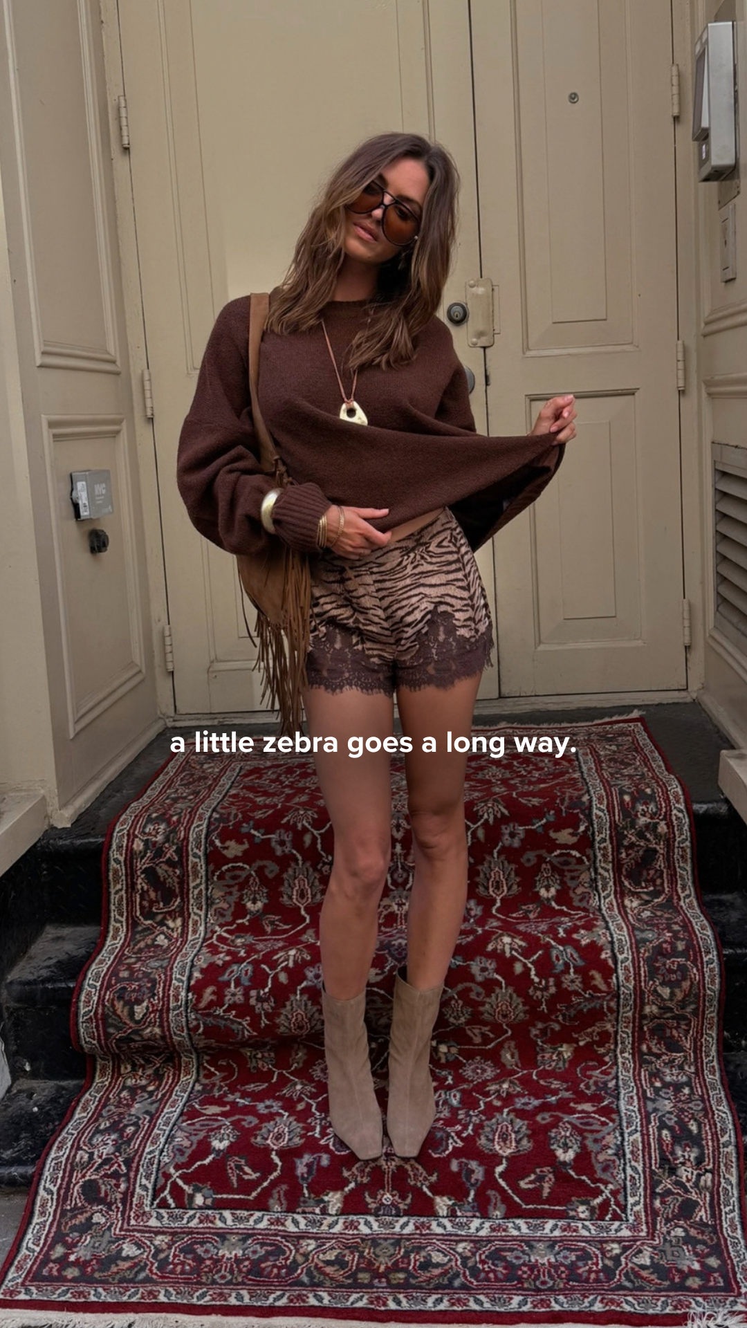 A woman stands on a red carpeted staircase in front of a door. She is wearing a brown sweater and patterned shorts. The text on the image reads "a little zebra goes a long way." The woman is looking down and holding the hem of her sweater.