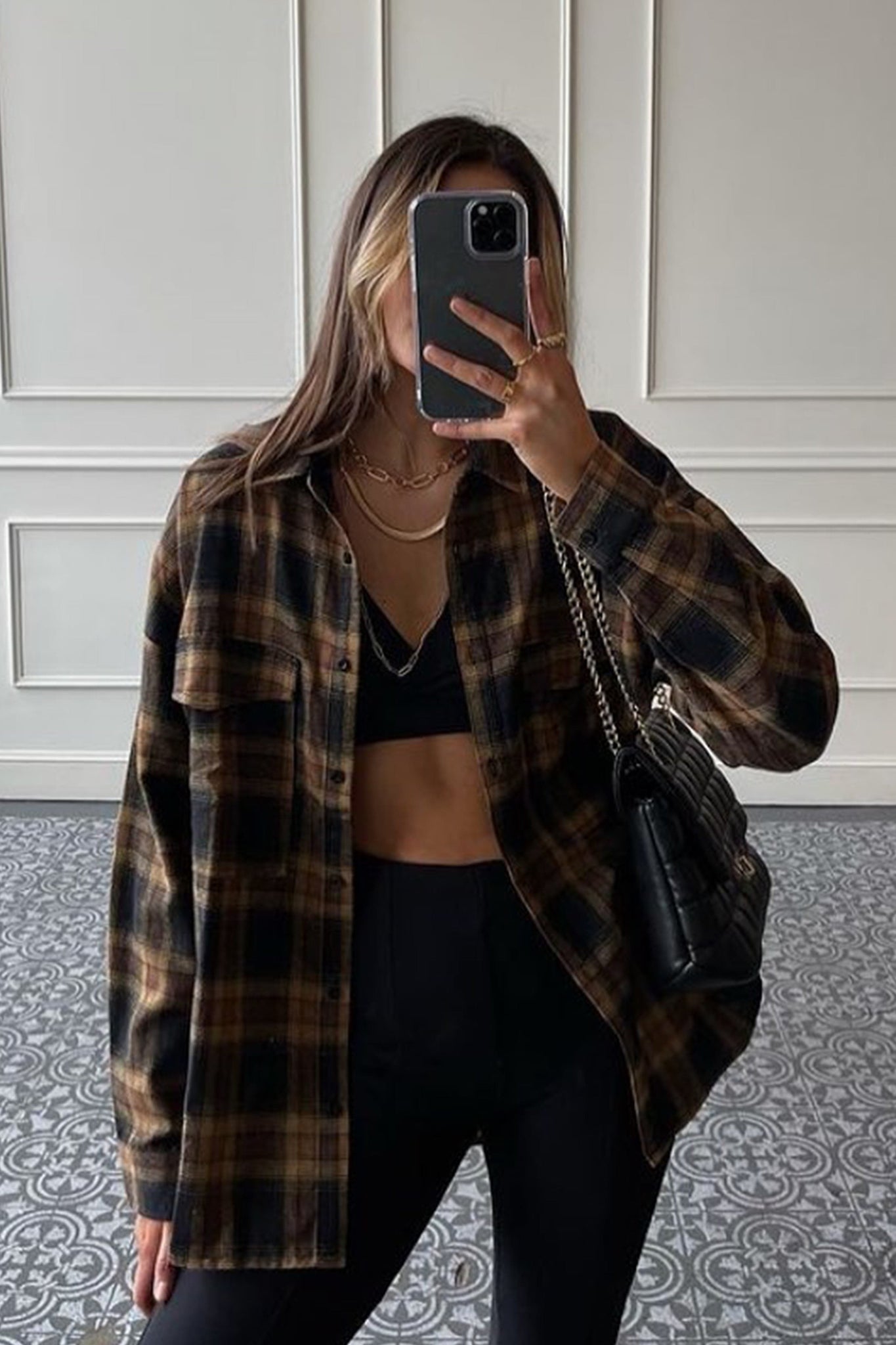 A woman takes a selfie in a mirror, wearing a brown and black plaid oversized shirt unbuttoned over a black bralette and black leggings. She is accessorized with a gold chain necklace and rings on her fingers, and carries a black quilted handbag over her shoulder. The text overlay reads: "The Internet’s Favorite Fashion Brand. Join the Tribe, and let’s make Memories."