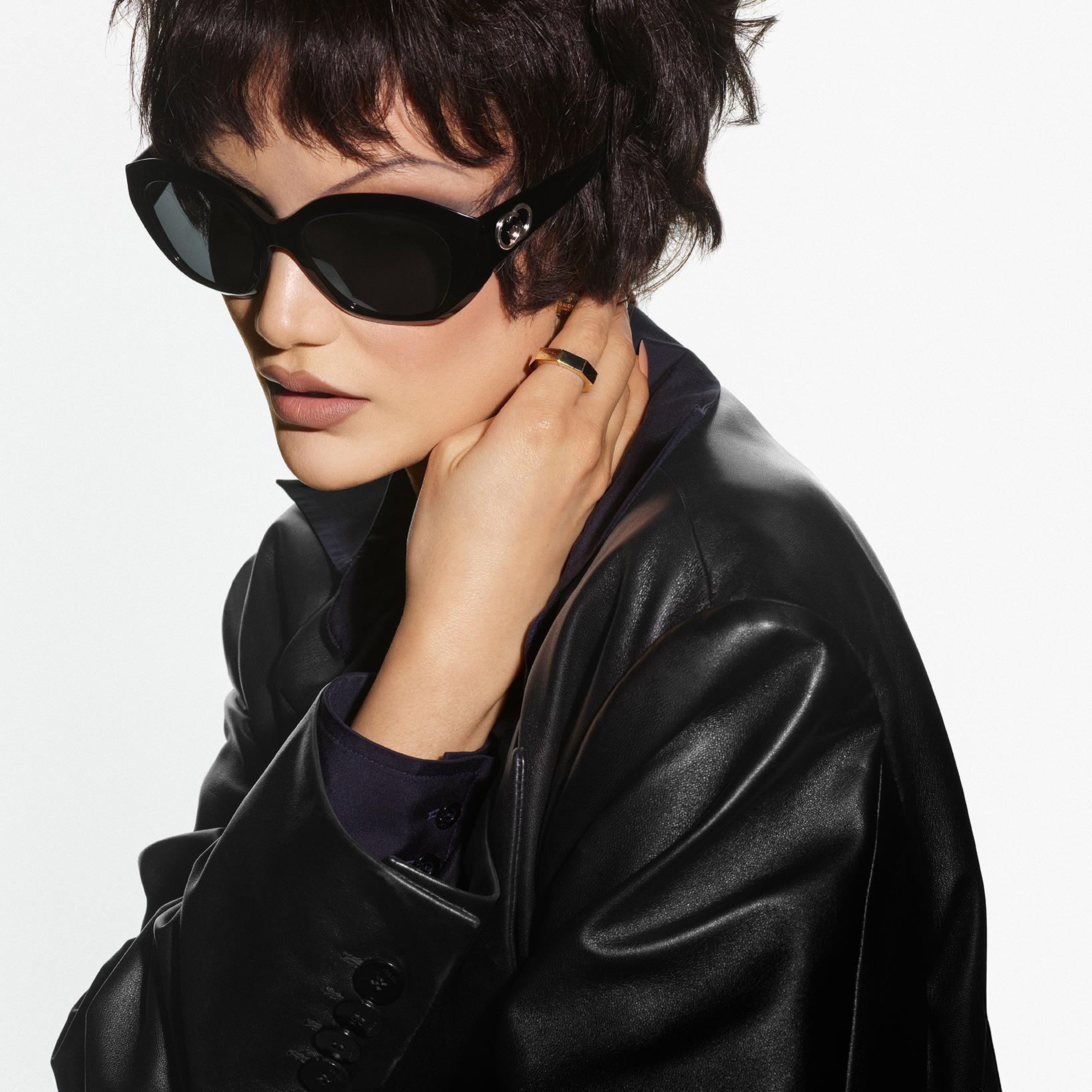 A close-up studio shot features a model wearing large, black, cat-eye sunglasses with gold accents on the temples. The model has short, dark, wavy hair and is wearing a black leather blazer over a dark collared shirt. They are touching their neck with one hand, revealing a gold ring on their index finger. The background is a plain white, creating a minimalist and high-fashion aesthetic. The image focuses on the model's face and upper body, highlighting the sunglasses and the overall chic style.