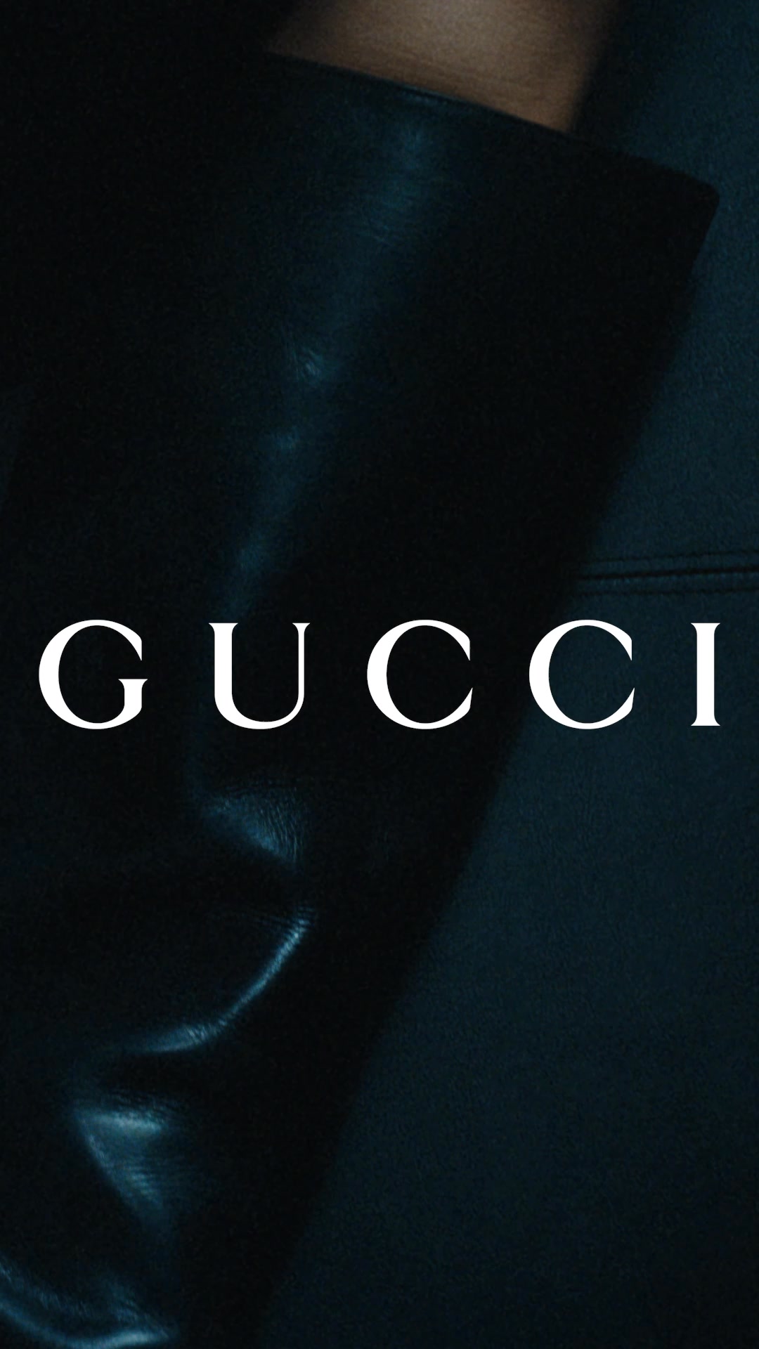 A close-up, dark, and moody shot focuses on the texture of a black leather garment. The iconic Gucci logo is prominently displayed in large, white, serif font across the center of the image. The lighting creates dramatic shadows and highlights on the fabric, emphasizing its luxurious quality. There is no additional text or offer presented in the creative.