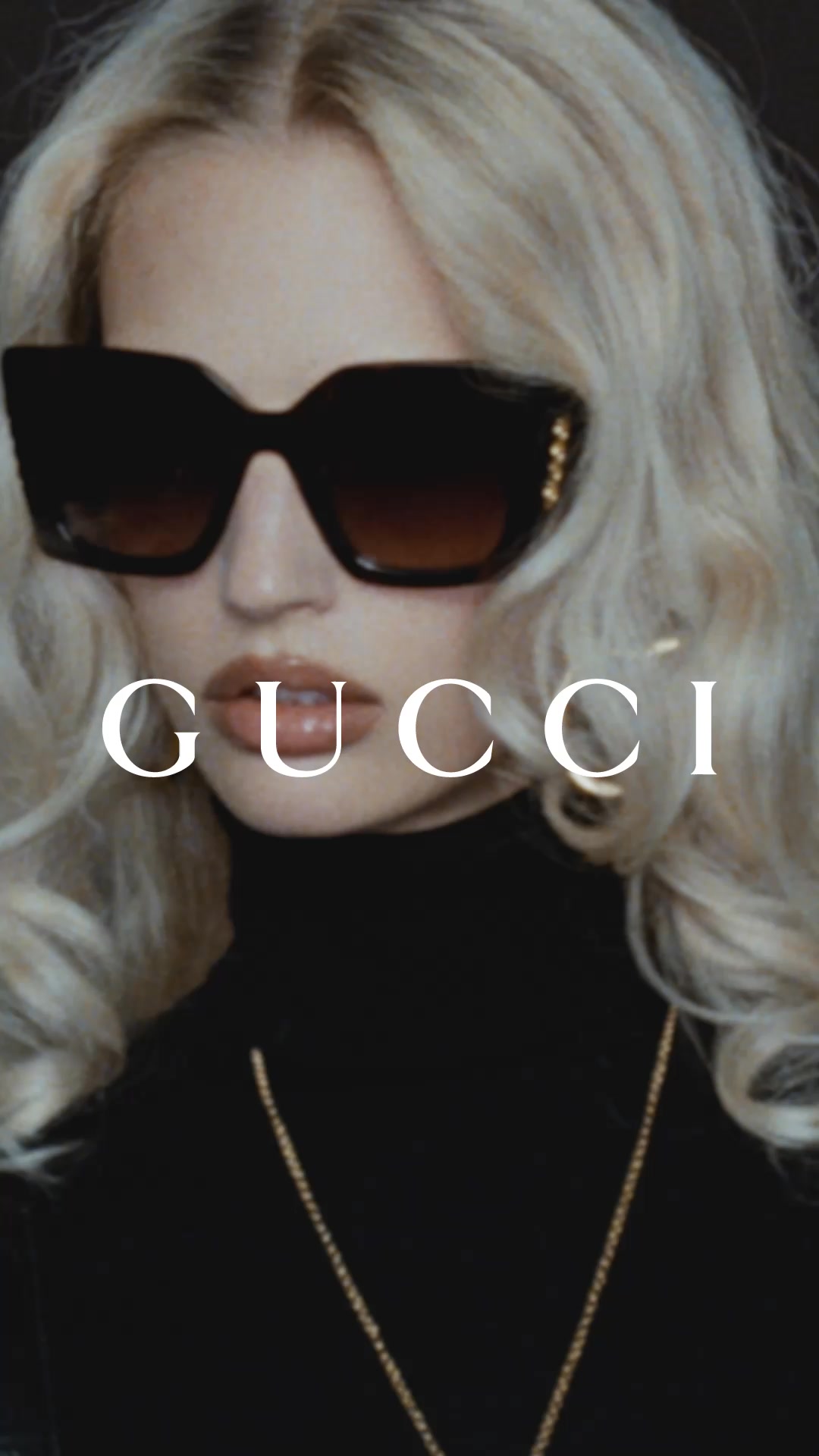A close-up shot of a woman with blonde hair, wearing large, dark Gucci sunglasses and a black turtleneck. White text that reads "GUCCI" is overlaid on the image, centered over her mouth. The overall aesthetic is glamorous and high-fashion, with a focus on the brand's iconic status and the contemporary style of the eyewear. The text directly addresses the viewer, introducing the new Gucci eyewear campaign.