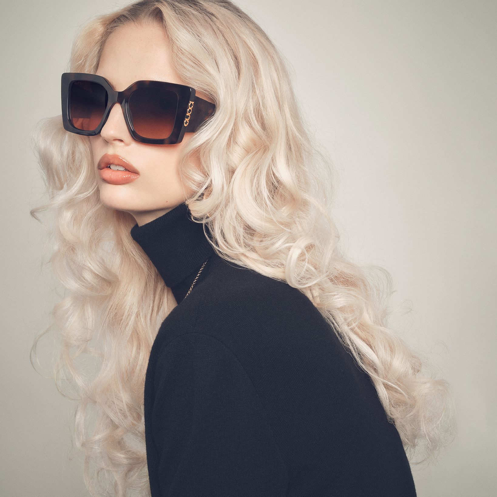 A close-up studio shot features a model with voluminous blonde hair, wearing large, tortoiseshell-patterned sunglasses with gold accents and a black turtleneck. The sunglasses are the main focus, with the Gucci logo visible on the side. The text overlay announces the bold silhouettes of the Spring-Summer 2026 Eyewear collection, highlighting the product's design features.