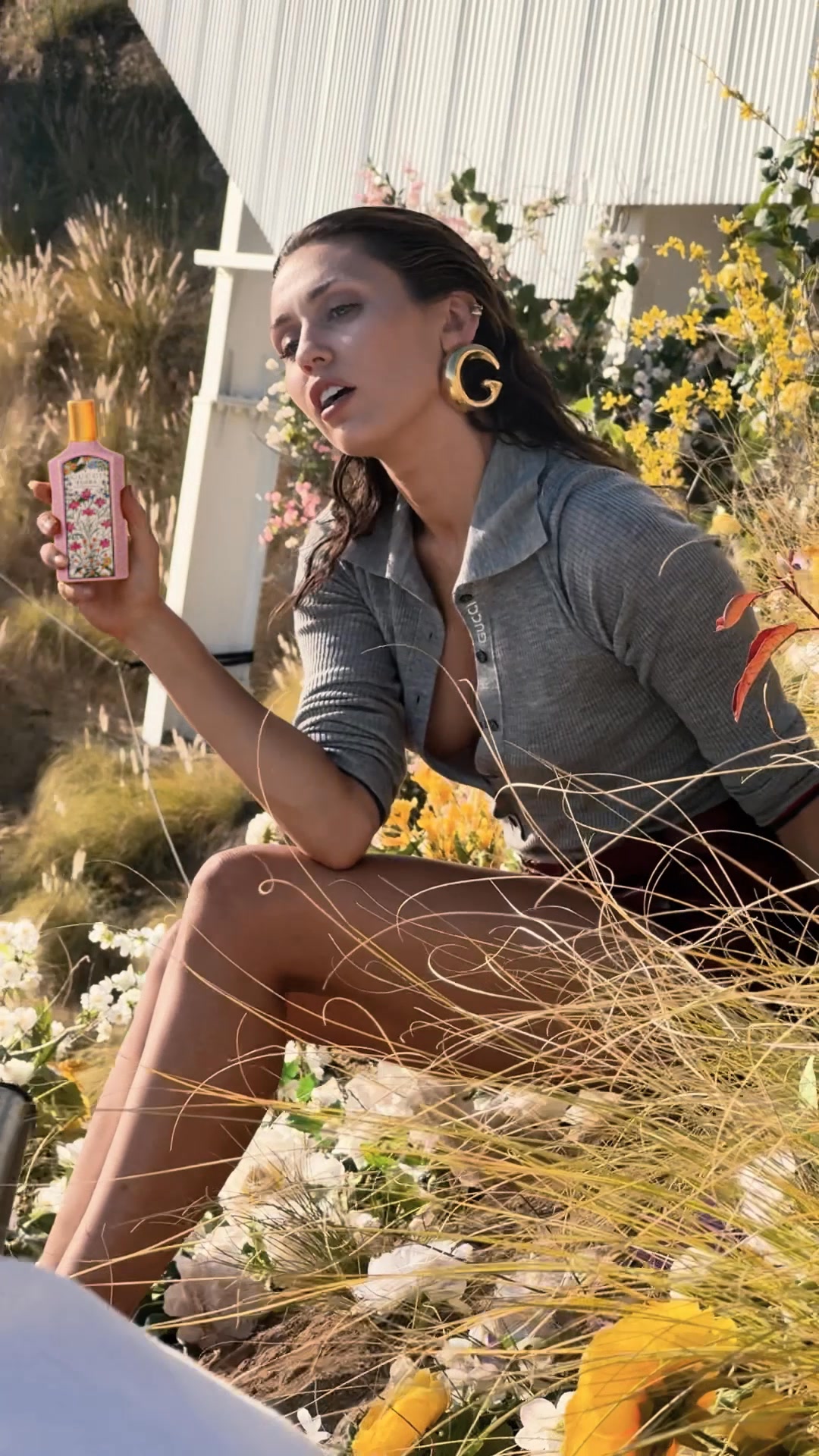A woman with wet hair, wearing a grey collared shirt and a dark skirt, sits outdoors surrounded by dried grasses and flowers. She holds up a pink bottle of Gucci Flora Gorgeous Gardenia Intense perfume. The text on the screen reads: "Miley Cyrus bringt ihre freie, unbeschwerte Art in Gucci Flora Gorgeous Gardenia Intense ein. #GucciBeauty #GucciFlora". The overall aesthetic is natural and bohemian.