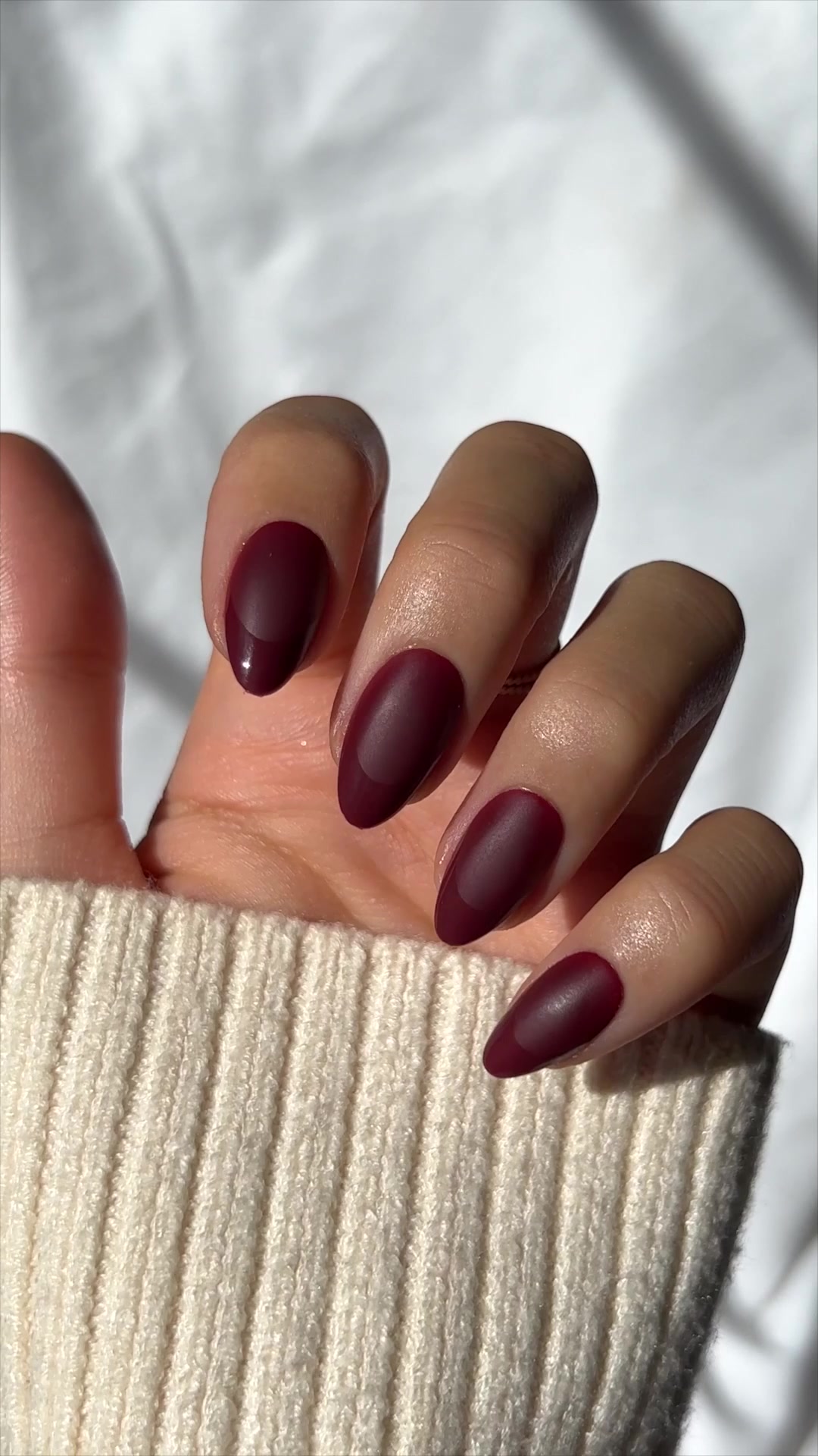 A close-up shot of a hand with burgundy, almond-shaped press-on nails is shown against a textured white background. The nails have a matte finish with a glossy crescent moon detail at the cuticle. The hand is partially covered by a cream-colored, ribbed sweater. On-screen text reads: "If you know, you know 😉". The image highlights the natural appearance and aesthetic of the press-on nails.
