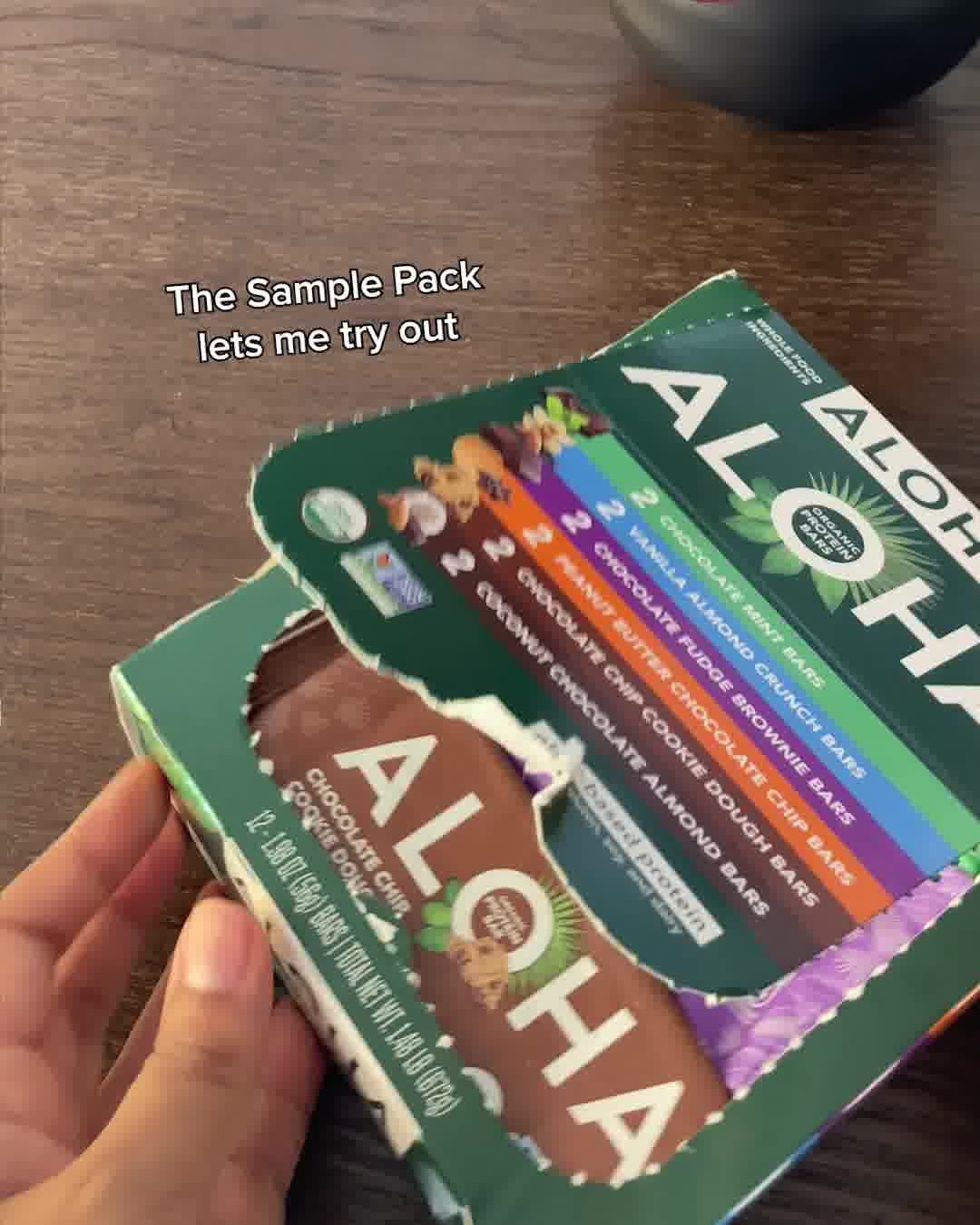A hand holds open a box of Aloha protein bars, revealing a variety of flavors inside. Text overlay reads "The Sample Pack lets me try out." The packaging is green and features the Aloha logo prominently. Several individual bars are visible, showcasing different flavor names and colors.
