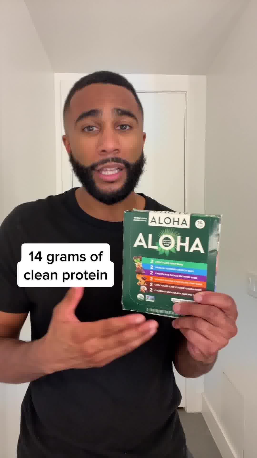 A man in a black t-shirt is holding a box of Aloha protein bars and gesturing with his other hand. On the left side of the screen, a white text box displays the words "14 grams of clean protein." The man is looking towards the camera and speaking. The product packaging is visible on the right side of the image, showing different flavors of the protein bars.