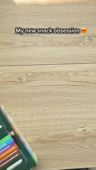 The creative features a wooden surface with the text "My new snack obsession 😍" overlaid. In the bottom left corner, a product with a green package and colorful edges is blurred and moving into the frame. The overall impression is of a casual, everyday moment where someone is excited about a new snack.