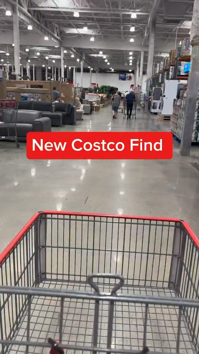 The video shows the interior of a Costco store from the perspective of someone pushing a shopping cart. In the foreground, the shopping cart is visible. In the middle ground, there are aisles with furniture and other products, and a few shoppers are walking around. A red banner with white text that reads "New Costco Find" is overlaid on the image. The overall impression is of a shopping trip and a discovery within the store.