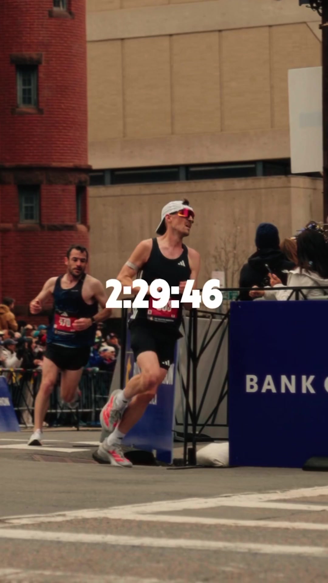 Two male runners are shown in profile, mid-race, with a digital timer overlaying the image reading "2:29:46". The runner in the foreground is wearing a black tank top, black shorts, a white cap, and sunglasses. The runner behind him is wearing a dark blue tank top and dark shorts. The background shows a blurred crowd and buildings. The creative is designed to highlight the performance and fueling strategies of elite marathon runners.