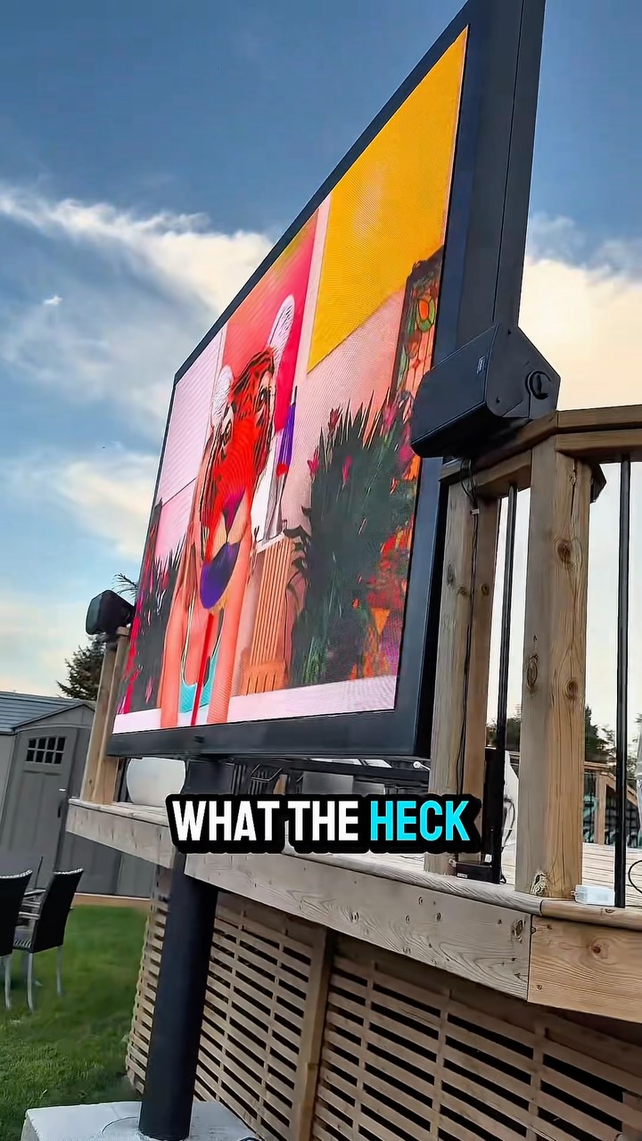 A large outdoor screen displays a colorful, abstract image that appears to be a collage of different visuals, including a tiger's face and floral patterns. The screen is mounted on a wooden deck structure. Bold white text with a blue outline reads "WHAT THE HECK" across the bottom of the screen. The background shows a cloudy sky and some greenery.