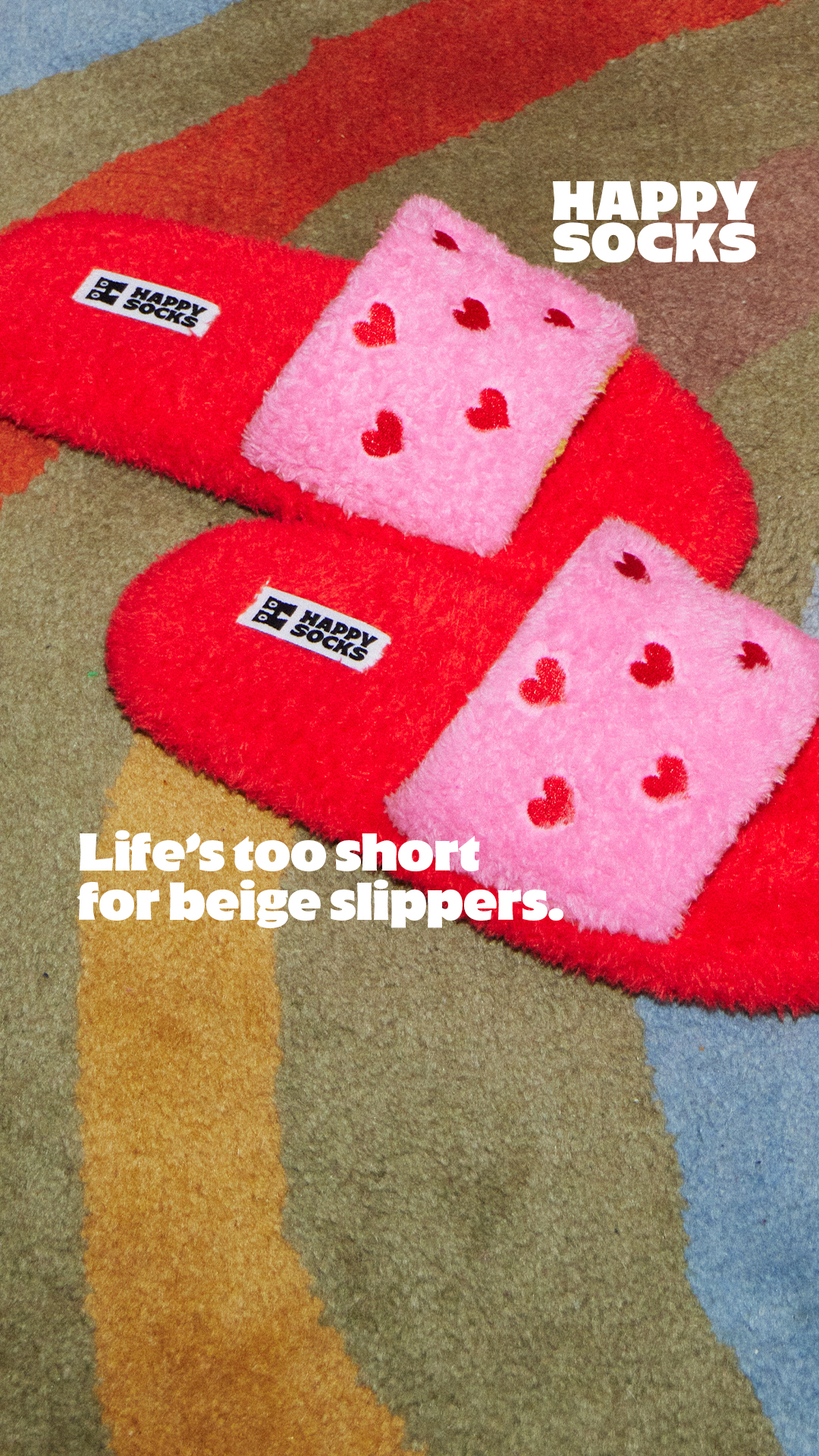A pair of bright red, fluffy slippers with pink fuzzy tops adorned with small red hearts are shown on a multi-colored striped rug. The Happy Socks logo is visible on a tag on each slipper. White text overlaid on the image reads "Life's too short for beige slippers." The overall impression is playful and emphasizes vibrant color choices over neutral ones.