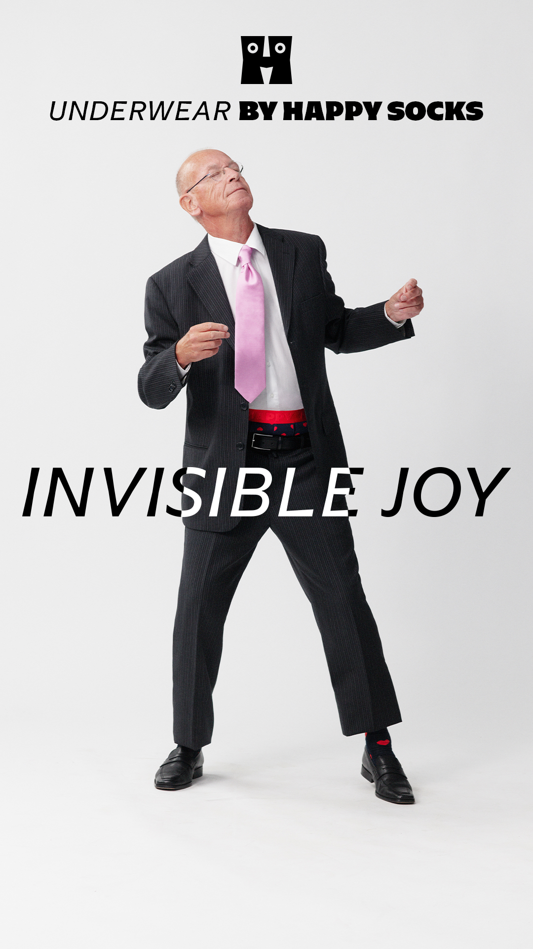 An older man in a dark pinstripe suit, white shirt, and pink tie is dancing with his arms outstretched. The text "UNDERWEAR BY HAPPY SOCKS" is displayed at the top of the image. Large, bold white text across the lower half of the image reads "INVISIBLE JOY". The man is wearing red and blue polka dot boxers, which are partially visible above his waistband. The overall tone is playful and energetic, suggesting that the underwear brings happiness.