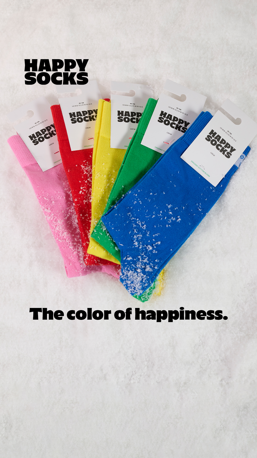A collection of five pairs of Happy Socks in vibrant colors (pink, red, yellow, green, and blue) are arranged in a fanned-out pattern on a white, snowy surface. The socks are partially covered in white specks, resembling snow or frost. Each pair has a white cardboard tag with the "Happy Socks" logo. The text "HAPPY SOCKS" is prominently displayed in bold black letters at the top of the image, and the tagline "The color of happiness." is written in bold black letters at the bottom.