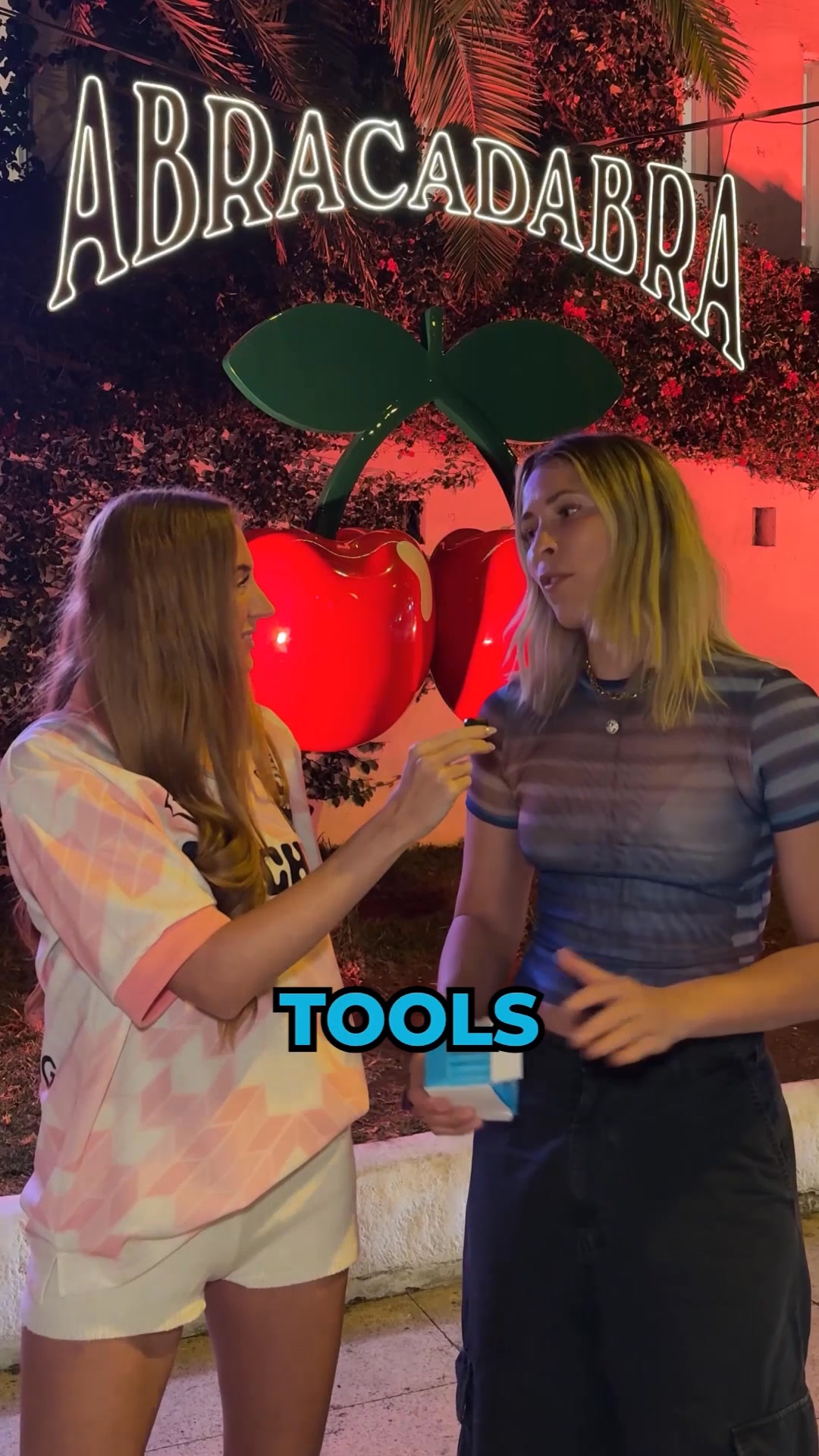 Two women are standing outside at night in front of a large neon sign that says "ABRACADABRA" and a giant cherry sculpture. The woman on the left is wearing a pink and white tie-dye shirt and white shorts. The woman on the right is wearing a blue and black striped mesh top and a dark skirt. The text "TOOLS" is overlaid in large blue letters on the bottom center of the image. The woman on the left is holding a small item up to the woman on the right, who is holding a small blue and white box.