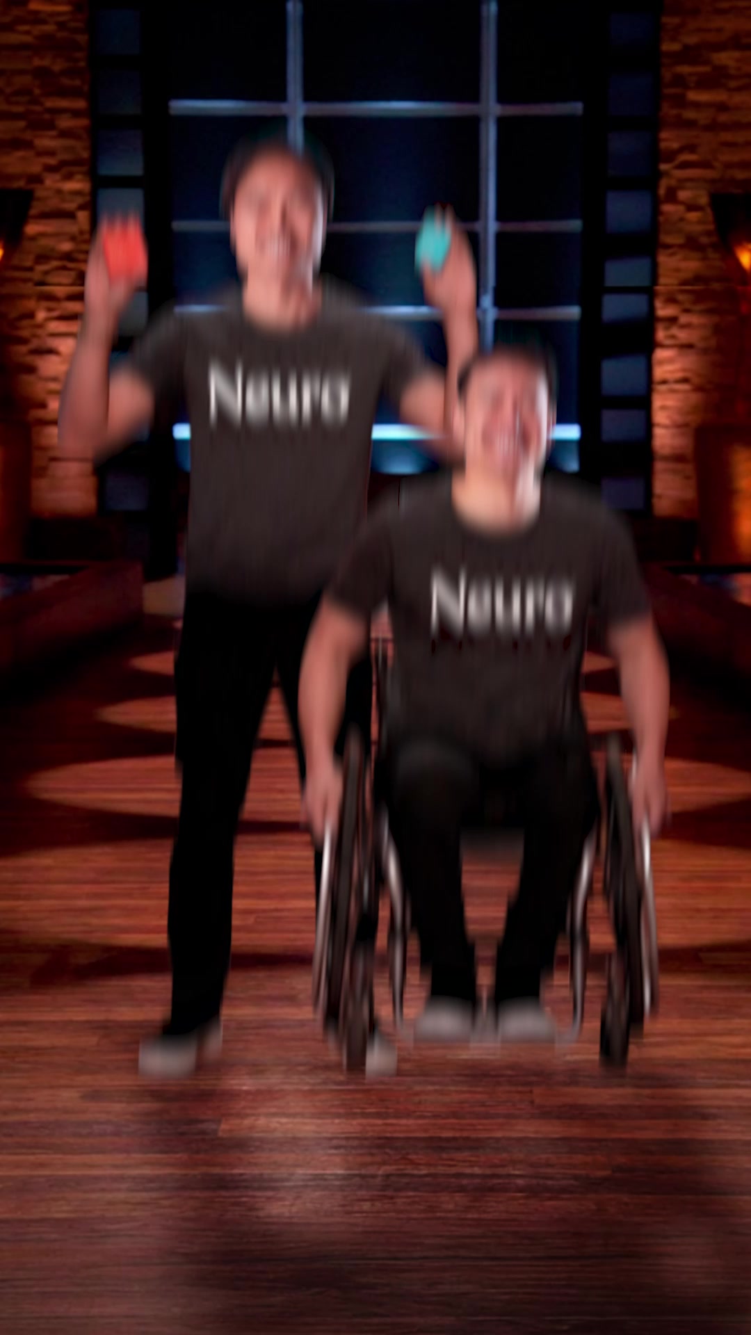 Two men are shown on a stage, one in a wheelchair and the other standing. Both are wearing dark t-shirts with the word "Neuro" in white text. The standing man is holding up two small, colorful objects, one in each hand. The man in the wheelchair is looking forward with a wide smile. The background appears to be a dimly lit stage with wooden paneling and grid-like windows. Text overlays the image, stating the brand's origin story and its benefits: "Neuro started as two friends with a mission to make energy cleaner, faster, and better for you and now, millions agree: We've Got Good Energy. 💚 Zero sugar ⚡️ Instant focus 🧠 Clean ingredients".