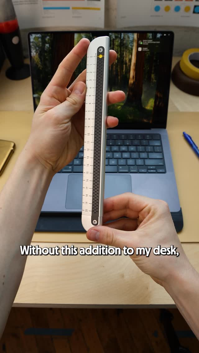 A person's hands hold up a white and grey ruler with black textured grips and measurement markings in centimeters. The ruler is positioned in front of a laptop screen displaying a forest scene. White text overlaid on the bottom of the image reads "Without this addition to my desk,". The creative focuses on the product and its utility in a desk setting.